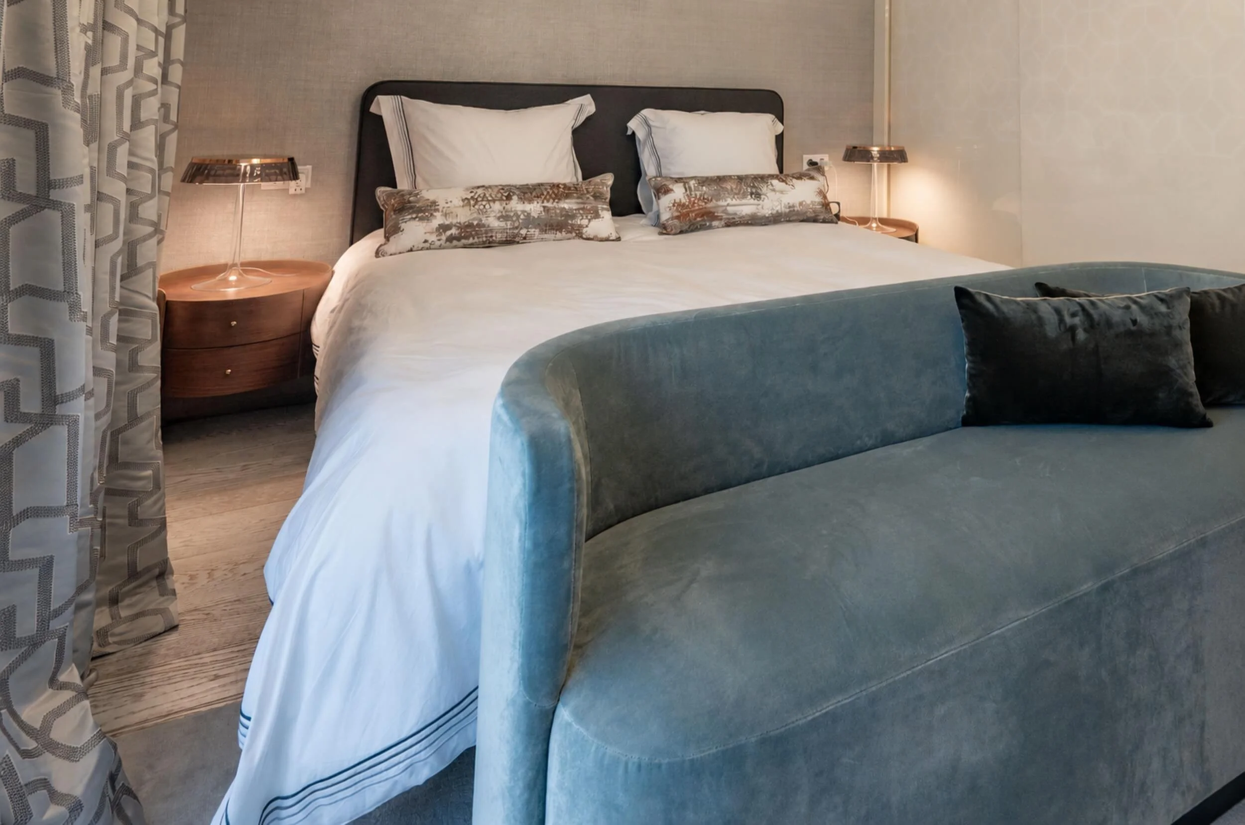 Detail des Main Bedrooms mit gepolstertem Bett, hellblauer Samtsitzbank, dekorativen Kissen und gerundeten Nachttischen aus Holz.