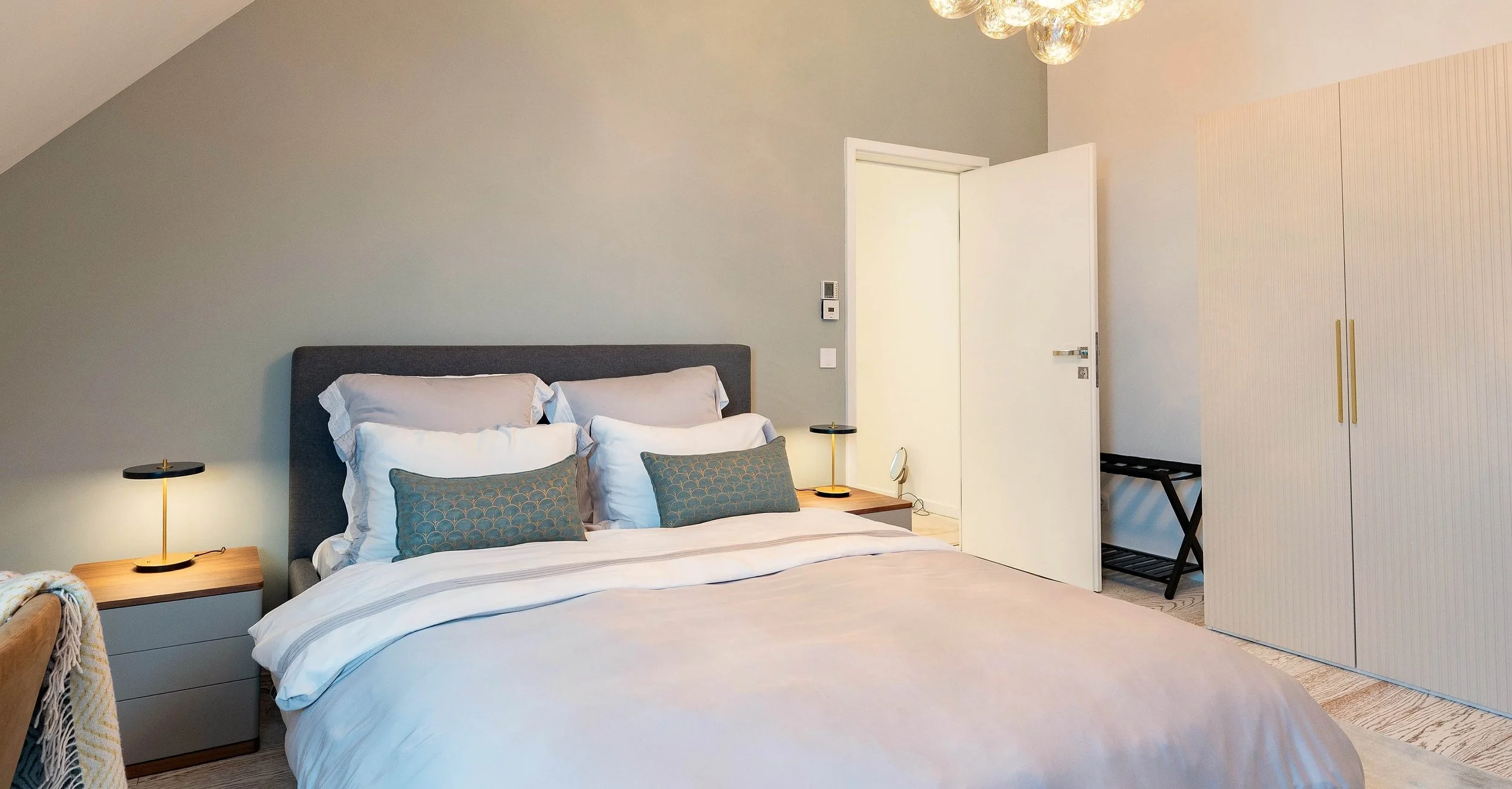 Gästezimmer mit gepolstertem Bett, Nachttischen in Grau und Holz, feiner Wandfarbe sowie hellem Schrank mit gerillten Fronten.