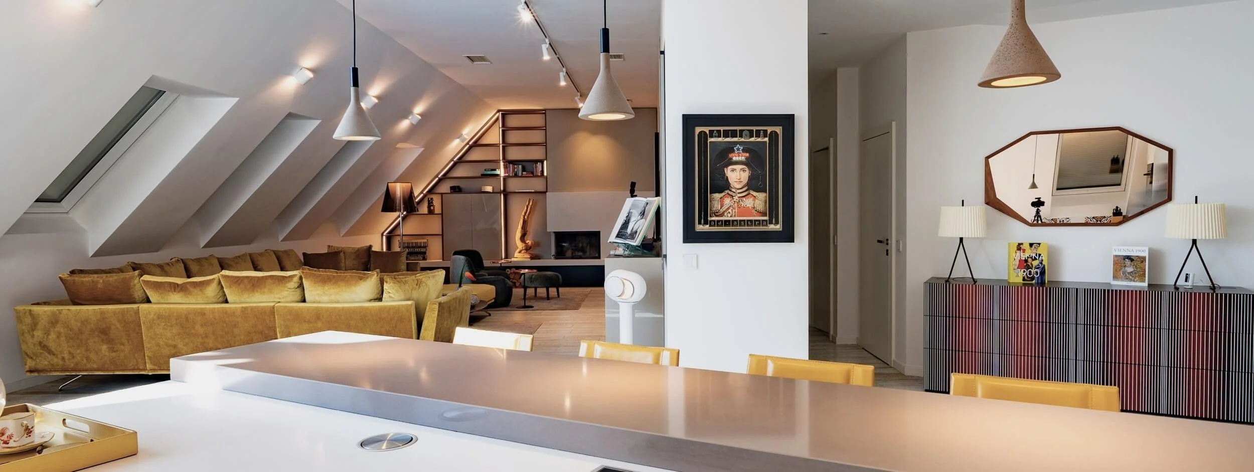 Offener Wohnbereich eines Rooftop Apartments in 1060 Wien mit Küche, senfgelber Sitzlandschaft, maßgefertigtem Kaminverbau, Kunst und Entrée.