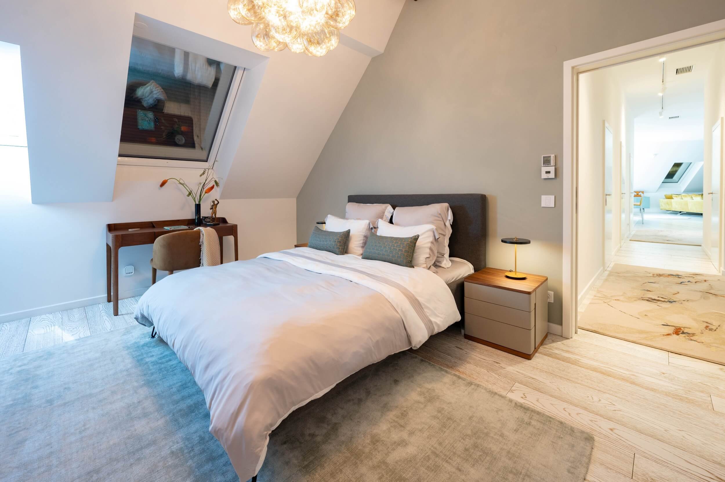 Helles Gästezimmer im Dachgeschoss mit gepolstertem Bett, integriertem Arbeitsplatz, Dachflächenfenster und zurückhaltender Farbgestaltung.