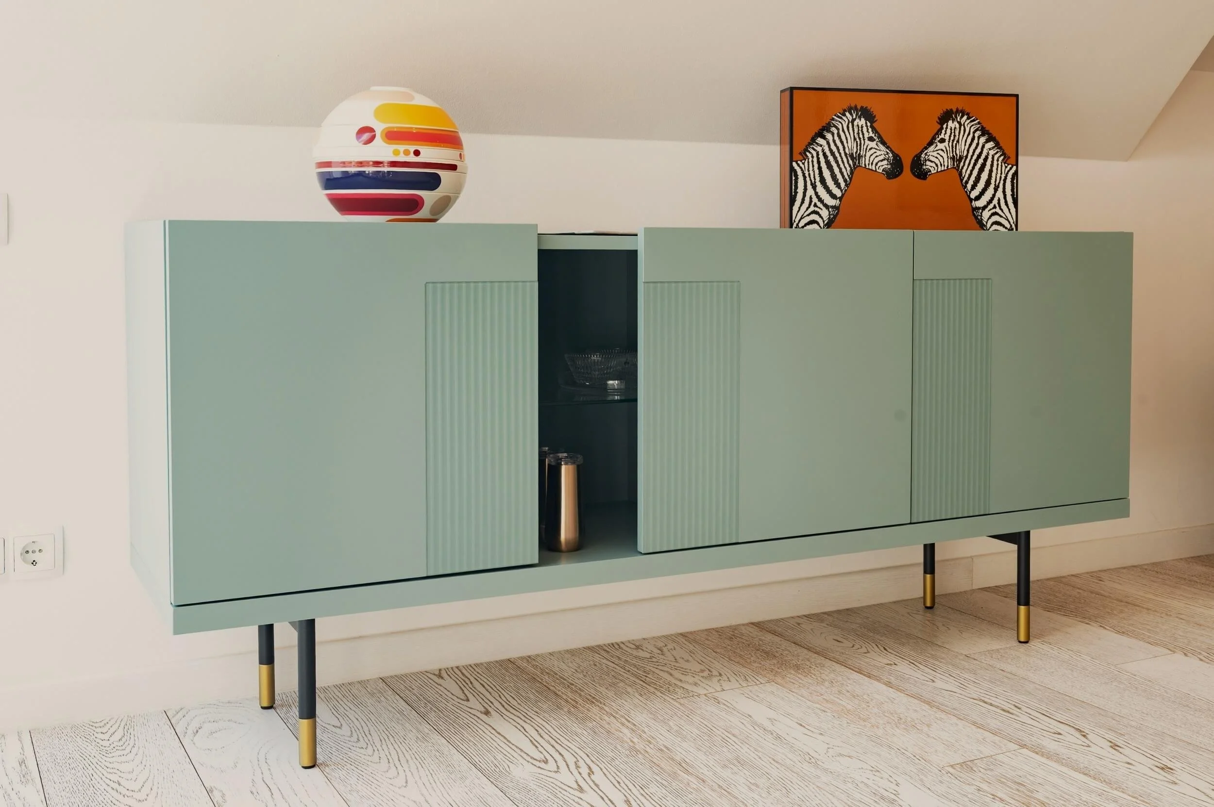 Detail eines farbigen Designobjekts auf dem petrolfarbenen Sideboard, ergänzt durch ein Kunstwerk mit Zebramotiv im Hintergrund.