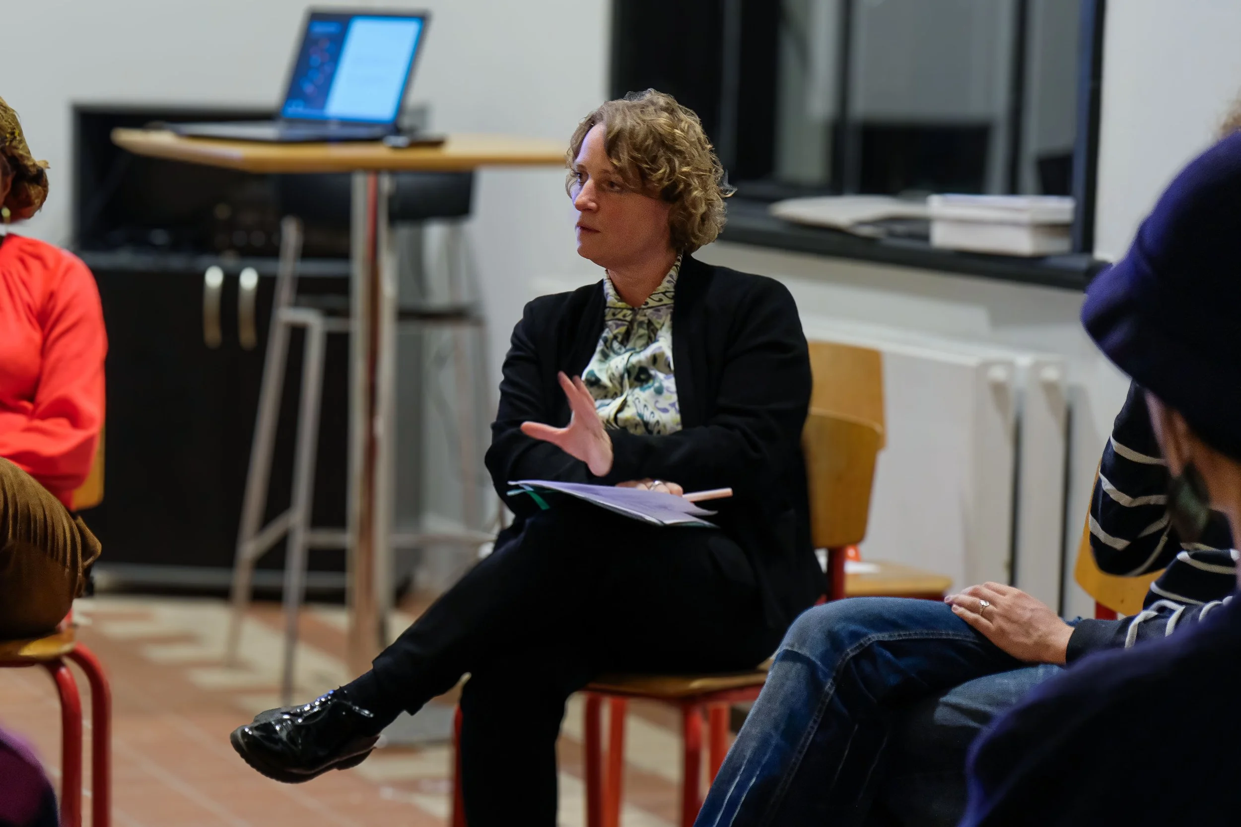 Henriette Hellstern as moderator sits on a chair in the art academy, talking during a discussion. She is holding papers and gesturing with her right hand, with other people visible around her. The topic is diversity.