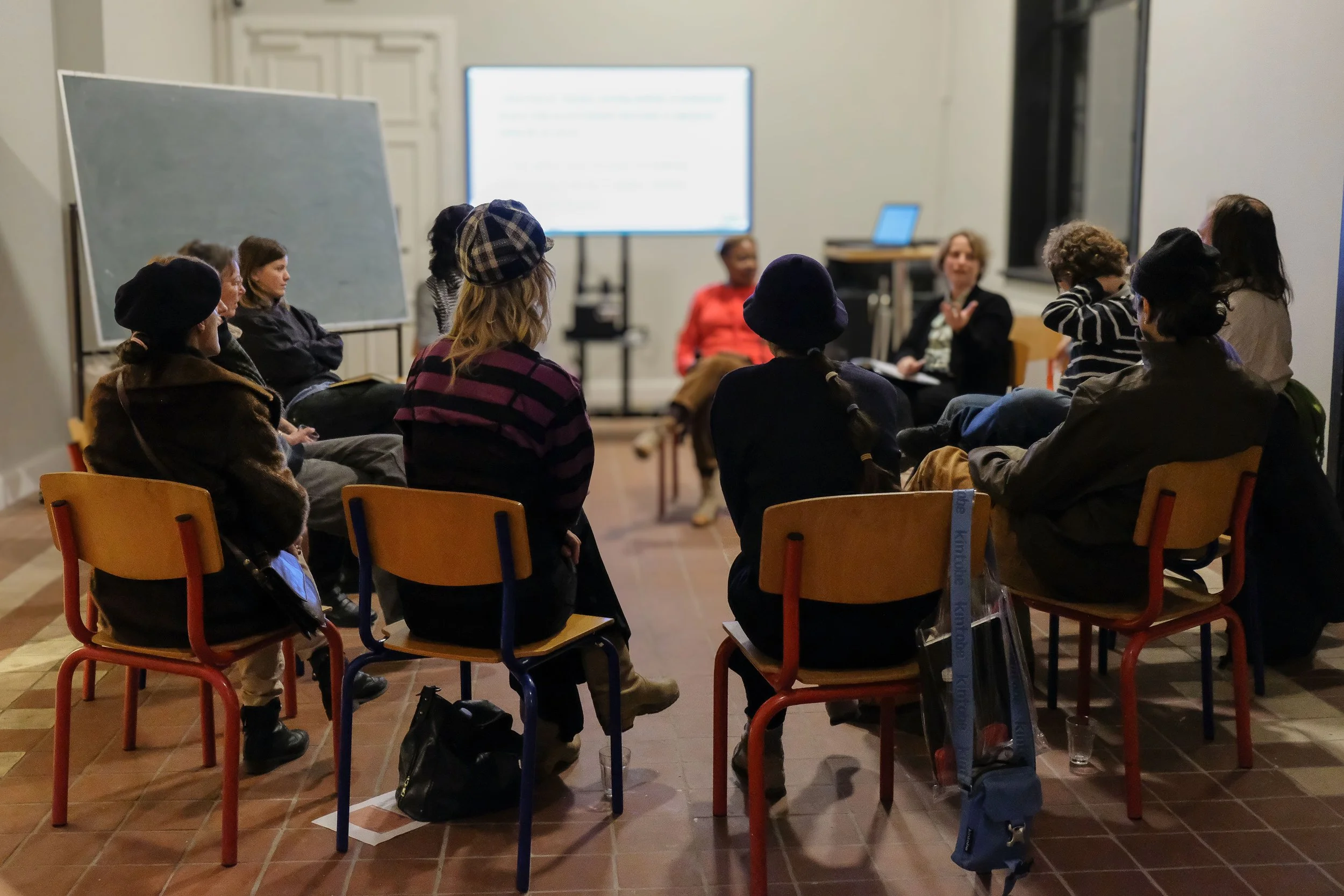 Art academy students sitting in a circle in a classroom-like setting, listening to Henriette Hellstern who is speaking. There are chairs arranged in a rectangular shape, and a large screen and a chalkboard are visible in the background.