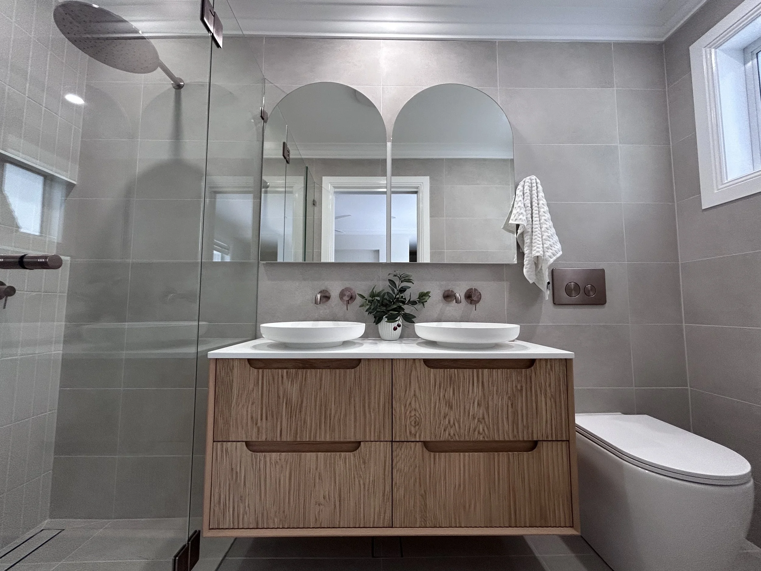 A modern bathroom with a double sink timber vanity, large mirror, towel, in wall cistern, and shower area, with gray tiled walls and a window.