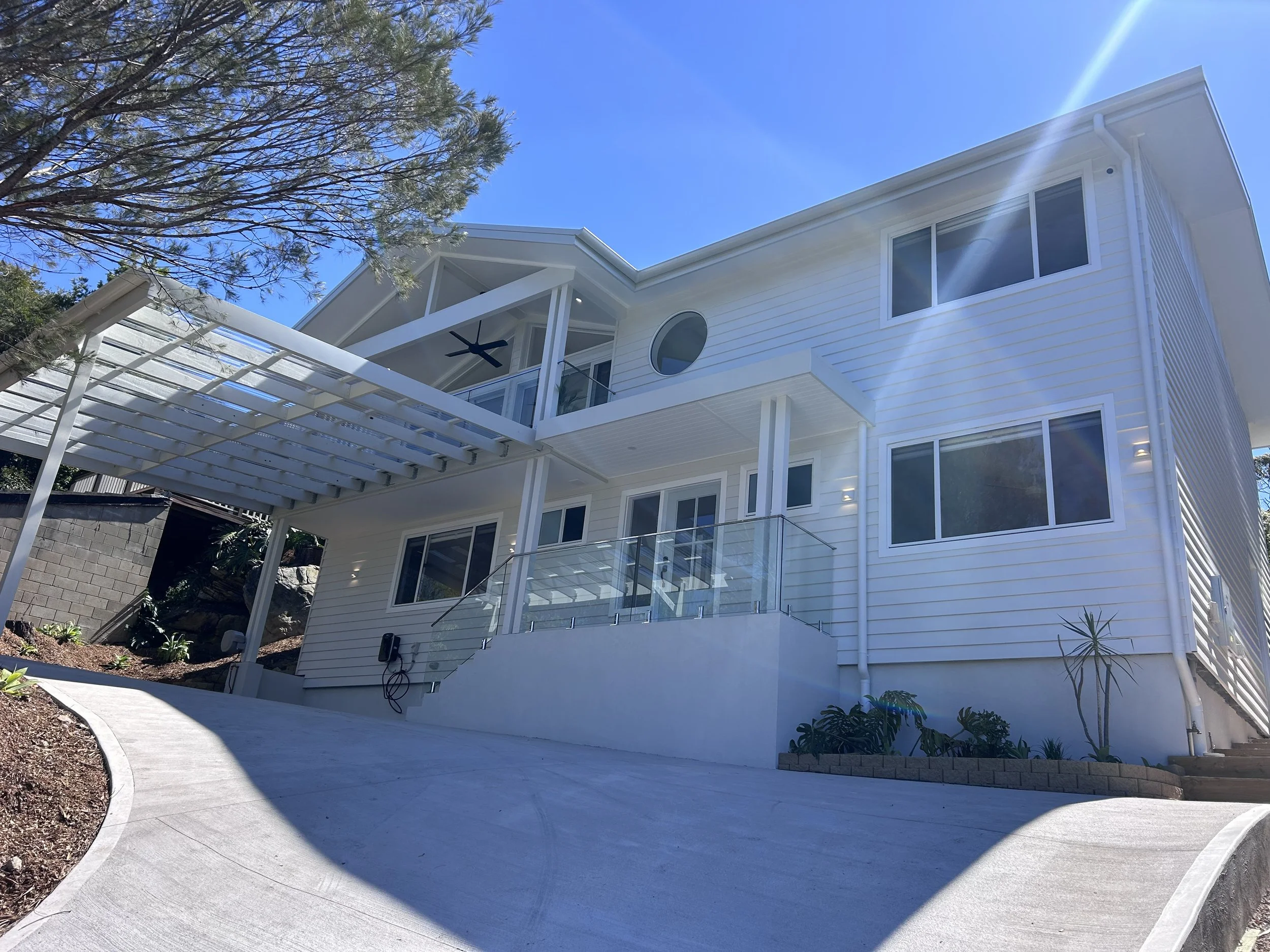 Modern coastal hamptons style house with multiple large windows, a balcony with glass railing, and a patio with stairs, surrounded by trees and a curved concrete pathway, under a clear blue sky.
