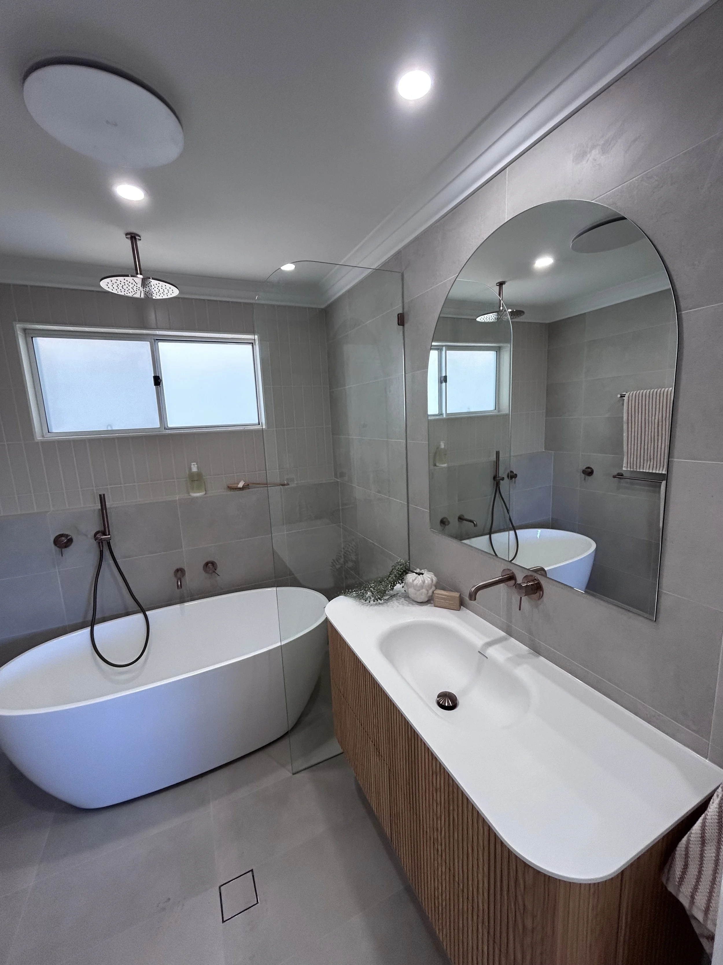 Modern bathroom with freestanding bathtub, large mirror, brushed copper champagne coloured tapware, timbergrain vanity, small window, beige tiled walls, and grey floor.