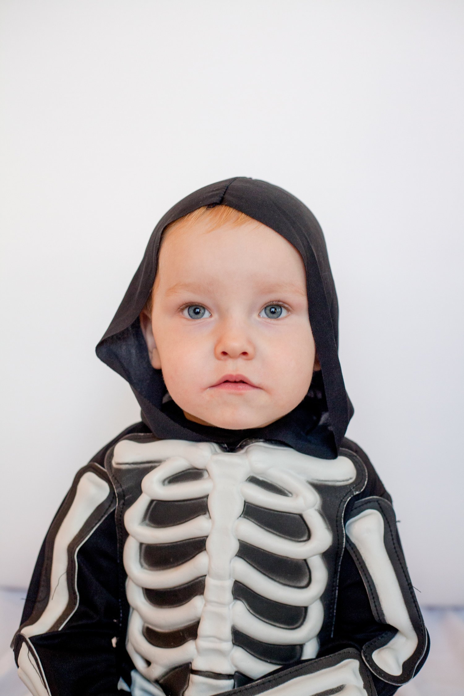 Young child dressed in a skeleton costume with a black hood and white bones, standing against a plain white background.