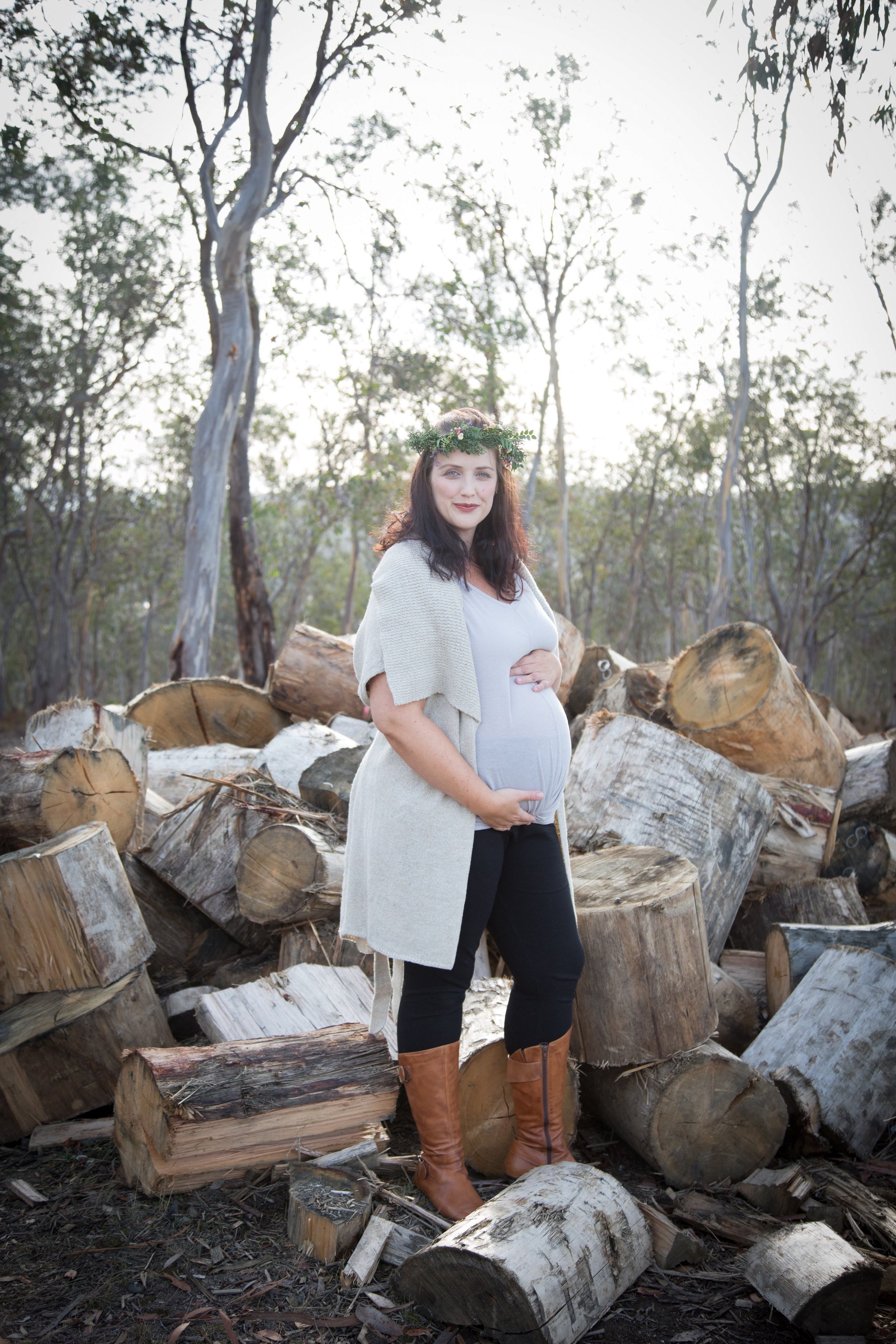 Pregnant woman with dark hair wearing a flower crown, light sweater, and black pants, standing outdoors in front of a pile of chopped logs and trees, with sunlight filtering through the woods.