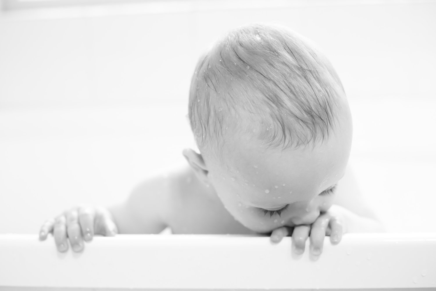 A baby with wet hair leaning on the edge of a bathtub, appearing to blow bubbles or interact with water, in a black and white photo.