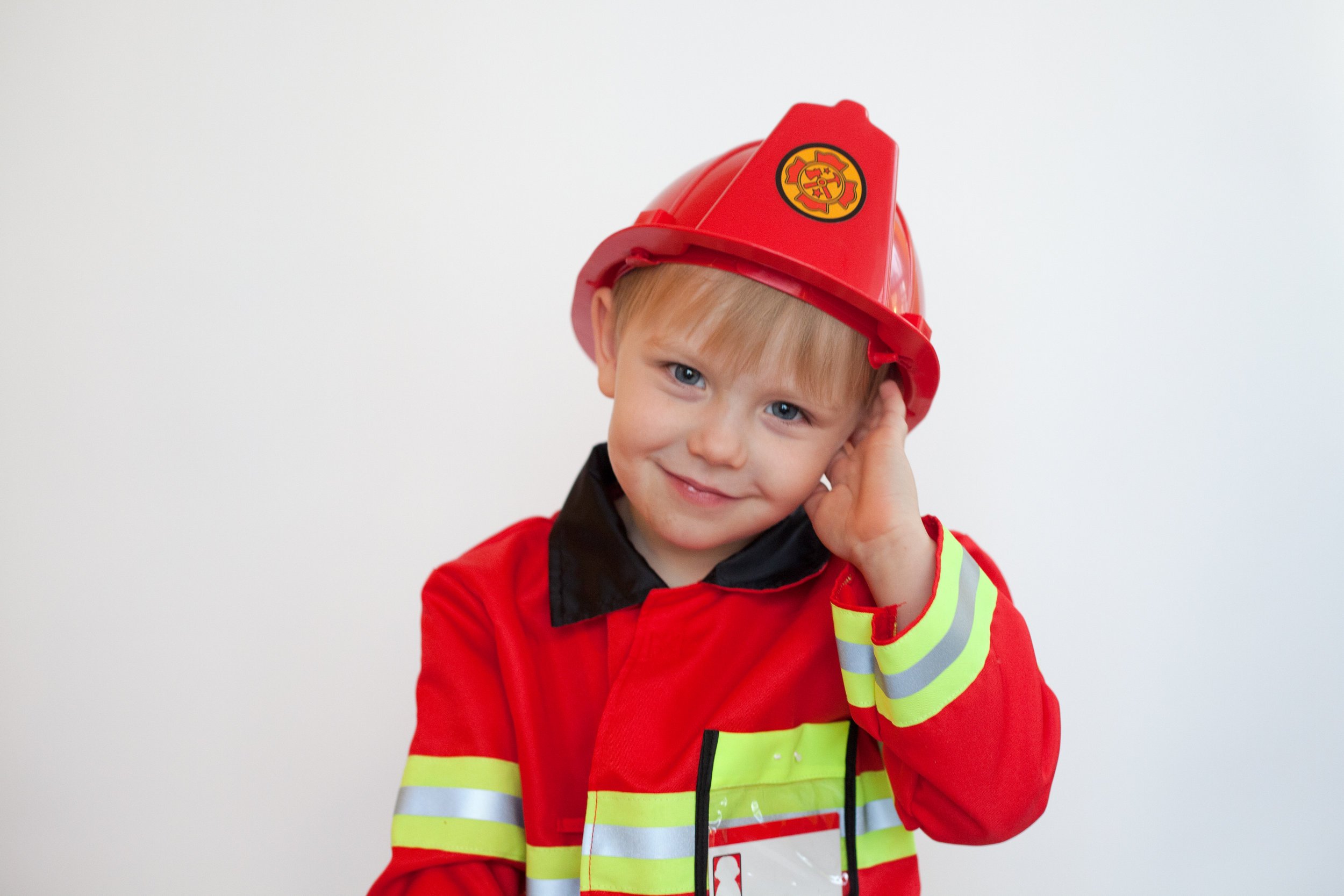 Young boy dressed as a firefighter wearing a red firefighter coat and a red helmet, standing against a plain white background before a fire station.