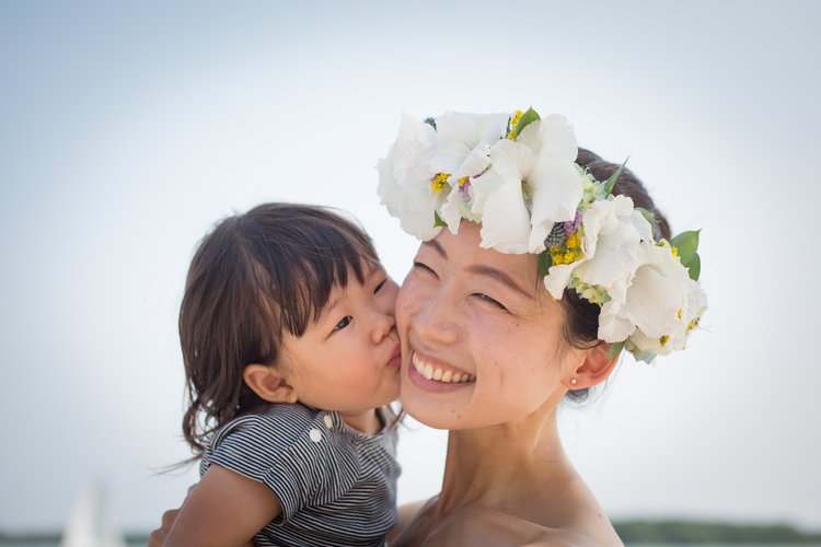 A young woman with a floral crown made of white flowers is smiling as a small child kisses her cheek.
