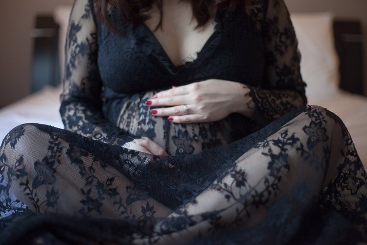 Woman wearing a black lace dress sitting on a bed with hands resting on her stomach.