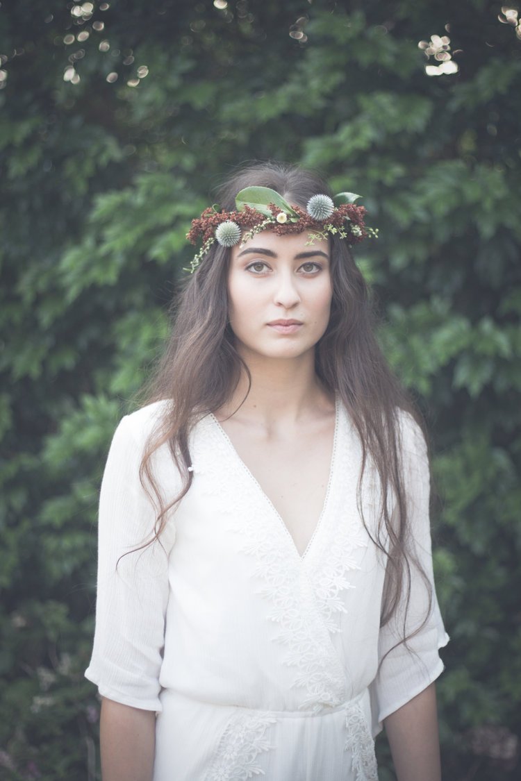 Young woman wearing a white embroidered dress and a flower crown, standing outdoors against a background of green foliage.