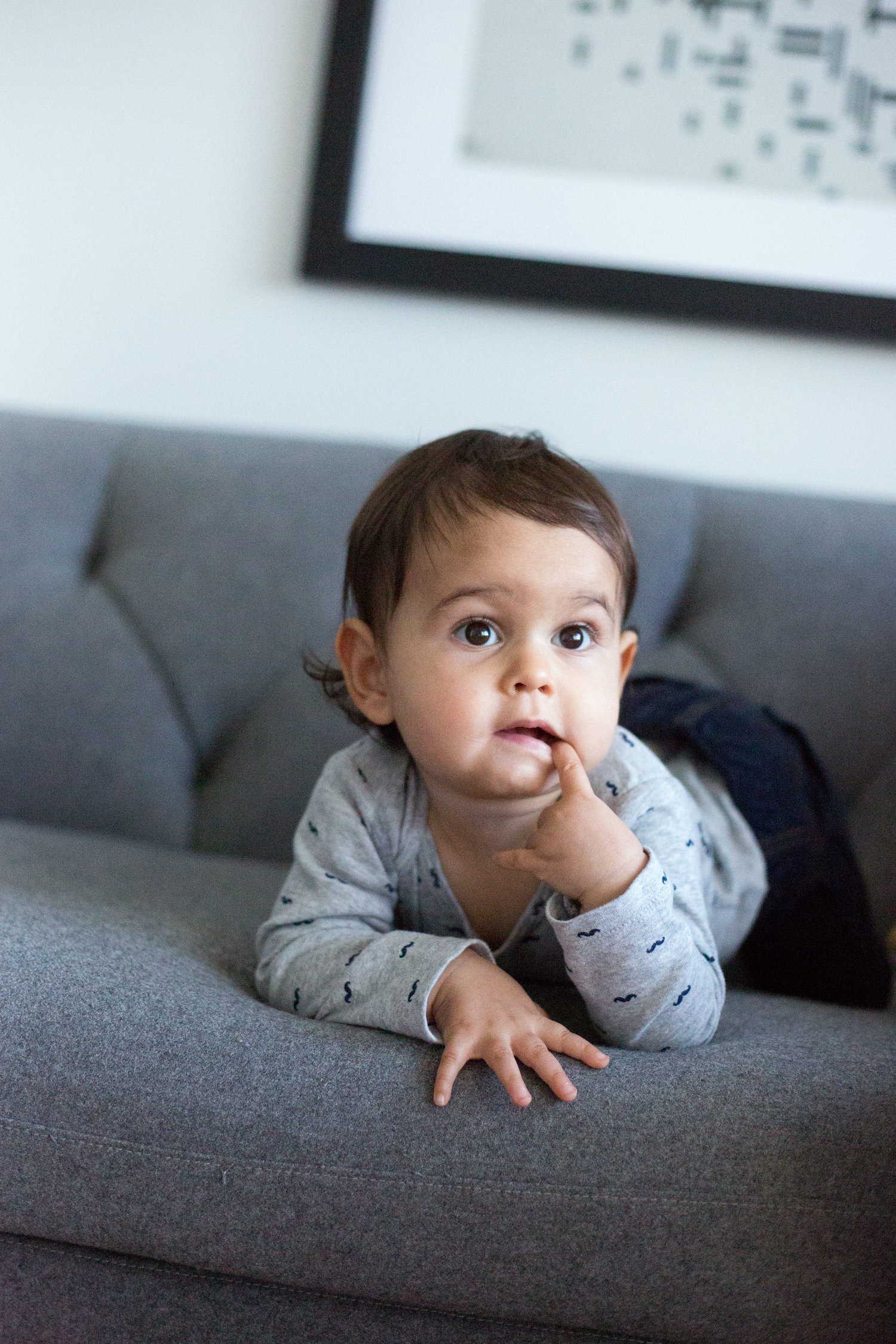 A young child with brown hair and big eyes, lying on a gray sofa with one finger in their mouth, in a room with a framed picture on the wall.