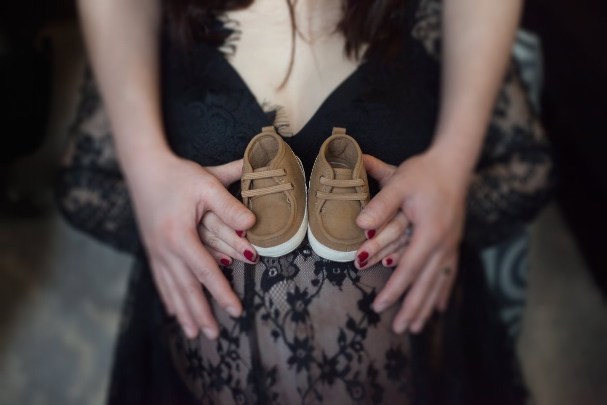 A woman in a black lace dress holding a pair of small tan baby shoes over her pregnant belly.