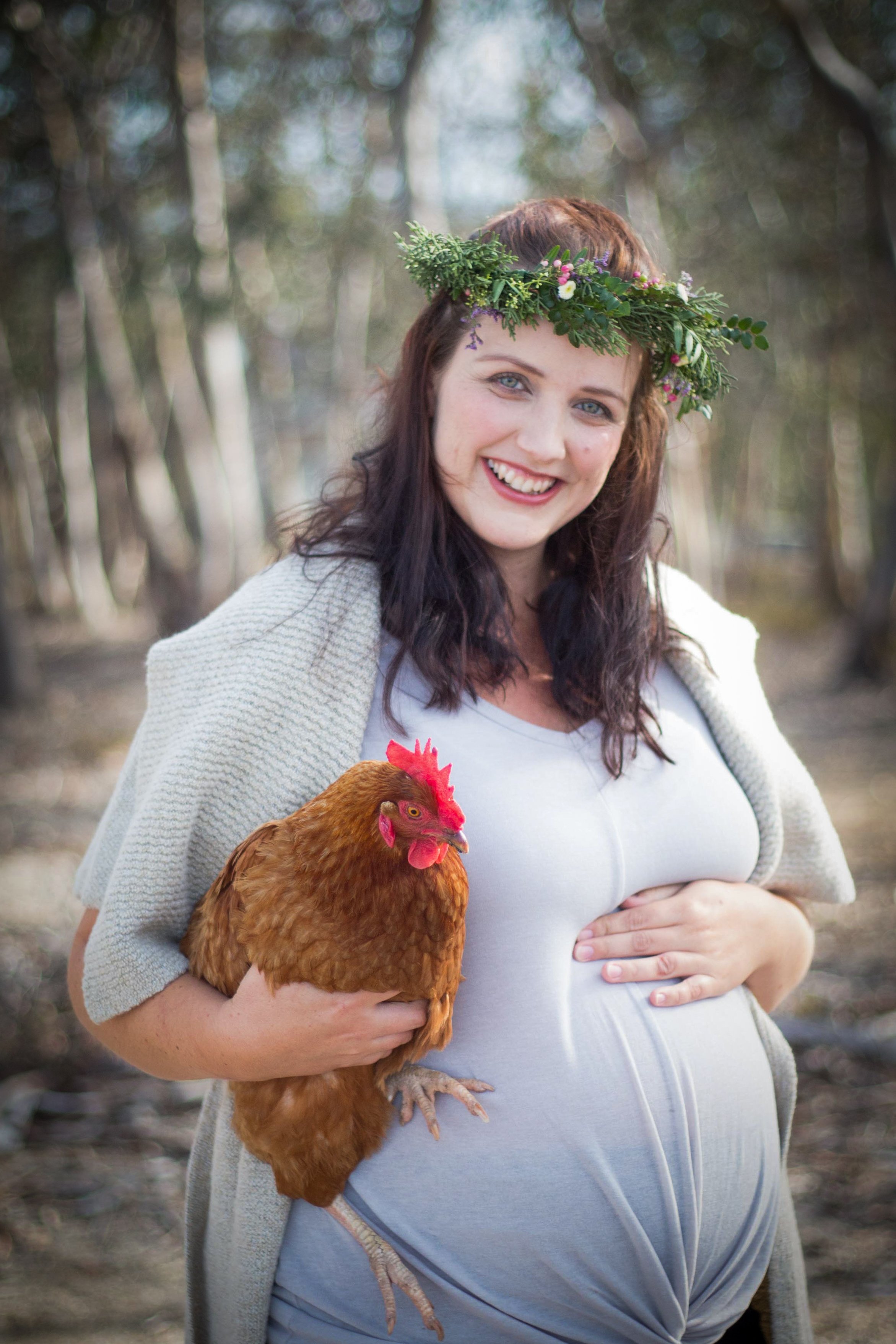 A pregnant woman smiling outdoors, wearing a flower crown, holding a chicken against a background of trees.