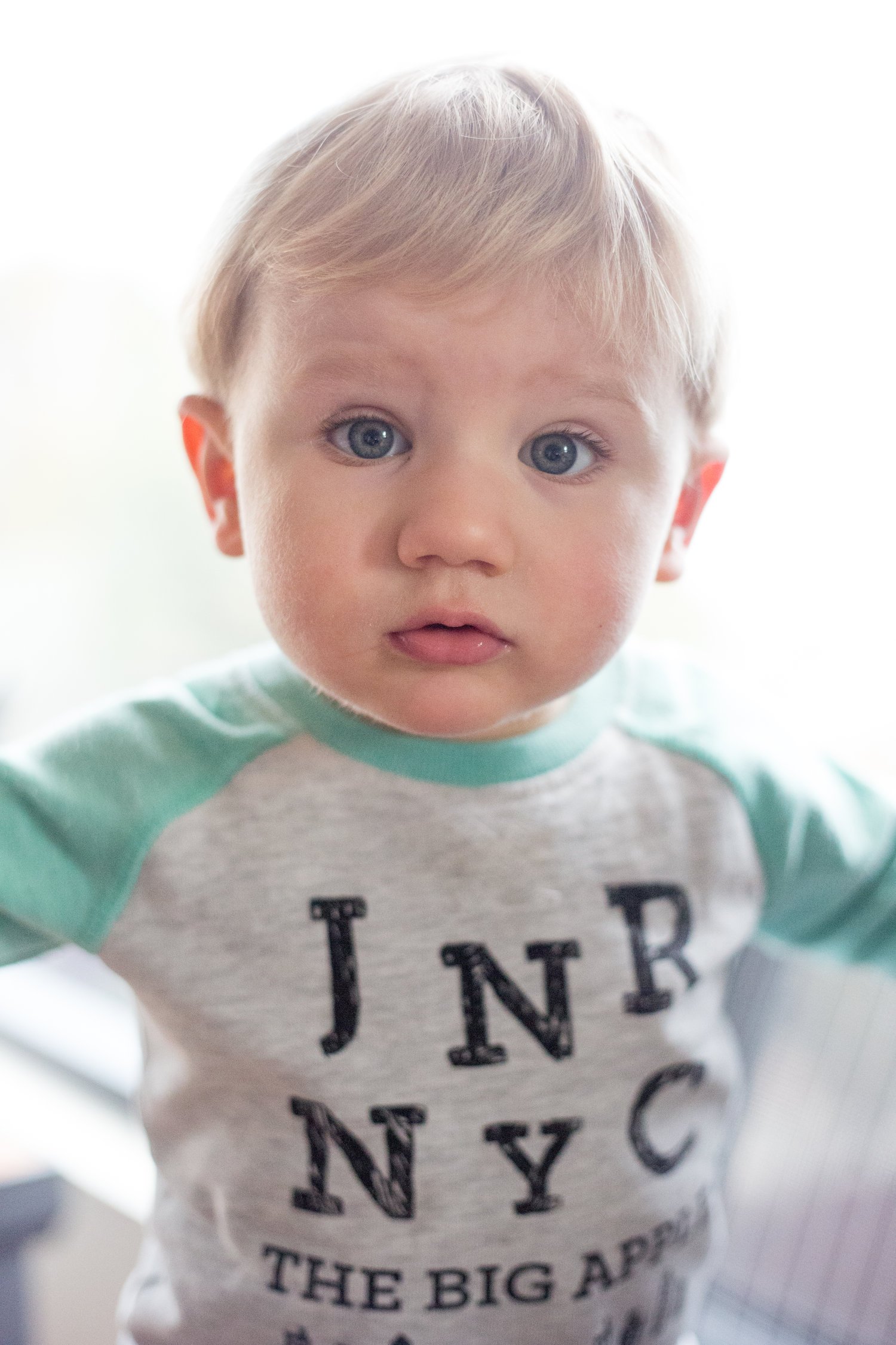 Close-up of a young boy with blond hair and blue eyes, wearing a gray shirt with green sleeves and black text.