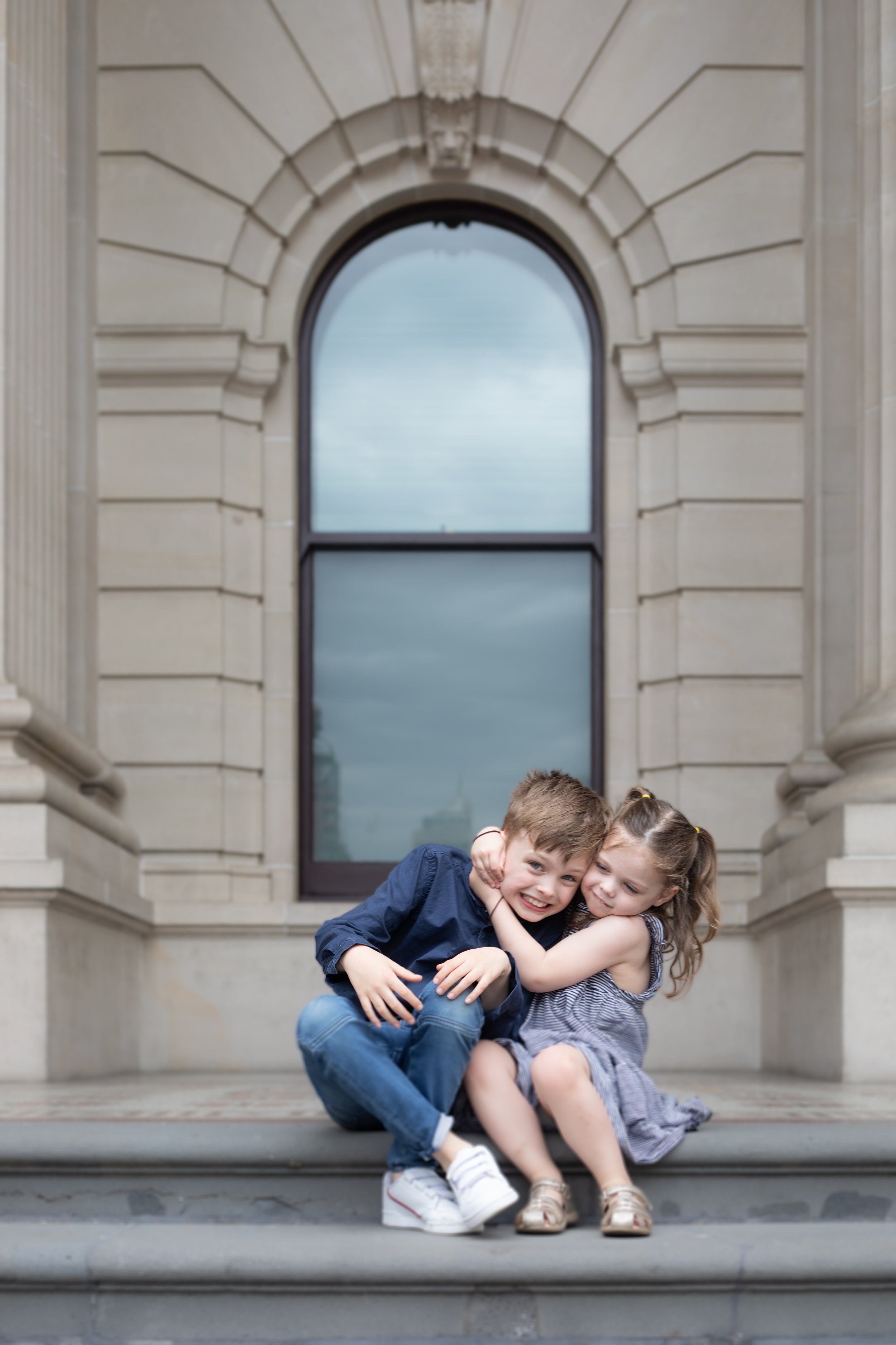 Two children, a boy and a girl, sitting on steps outside a building, hugging and smiling.