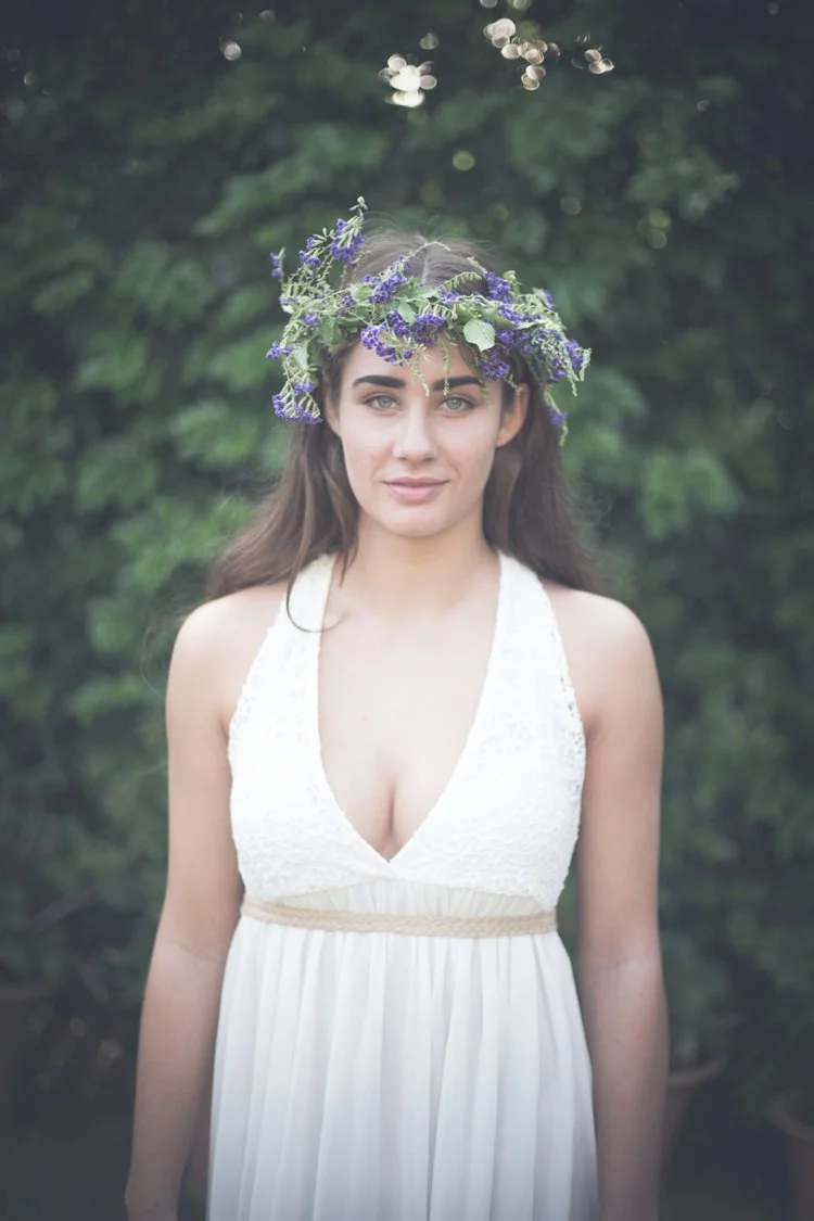 Young woman standing outdoors with a purple flower crown, wearing a sleeveless white dress, in front of a green leafy background.