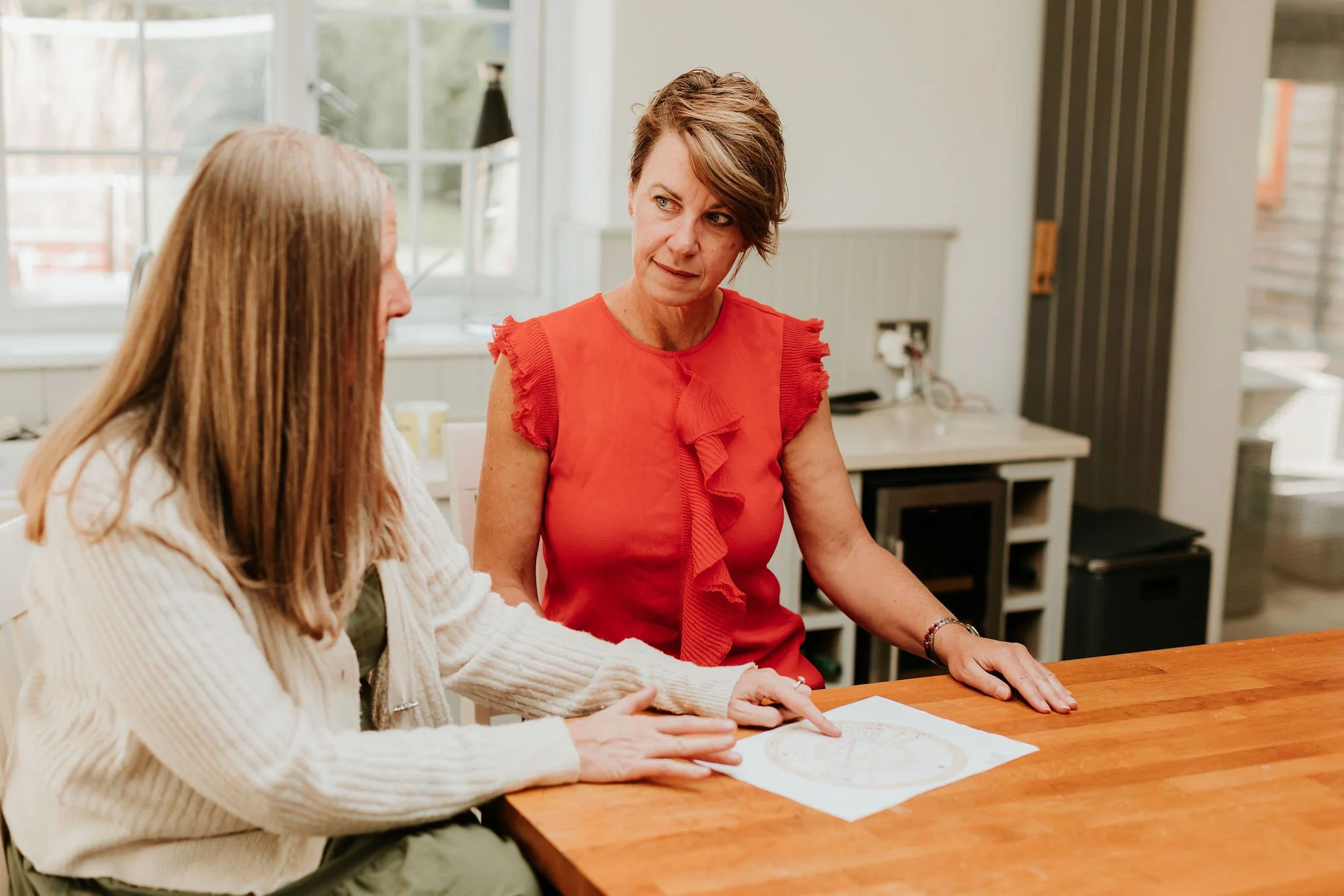 Two women sitting at a wooden table having a conversation, with a paper and diagram on the table, in a well-lit kitchen.