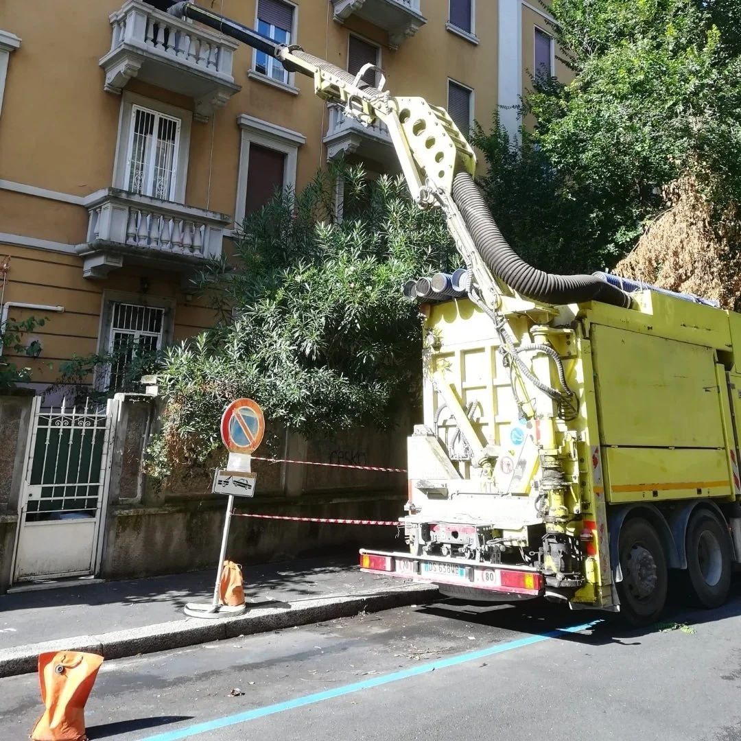 Una strada urbana con un camion per lavori di potatura o taglio alberi. Si vedono un albero e un edificio residenziale con balconi.