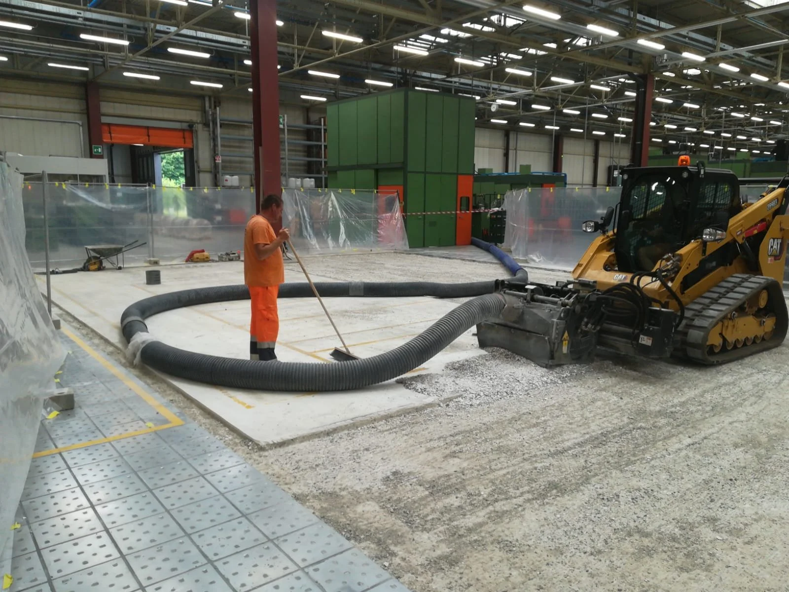 Operaio con vestiti arancioni che usa un tubo di aspirazione in un cantiere di costruzione all'interno di un capannone industriale. Sullo sfondo, una macchina bulldozer gialla, e strutture di stabilimento con pareti in lamiera e scaffalature verdi.