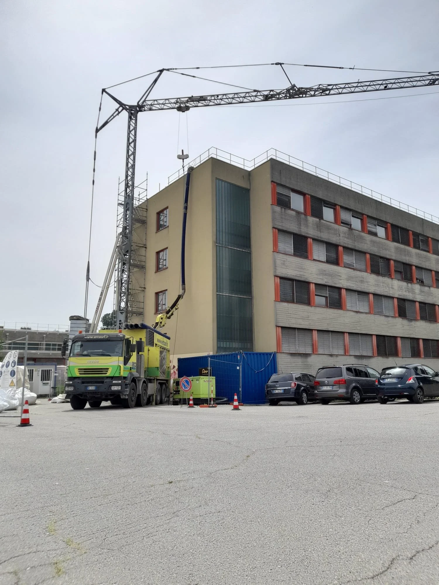 Cantiere edile con una gru sopra un edificio di quattro piani. Ci sono cinque auto parcheggiate e barriere di sicurezza con coni stradali