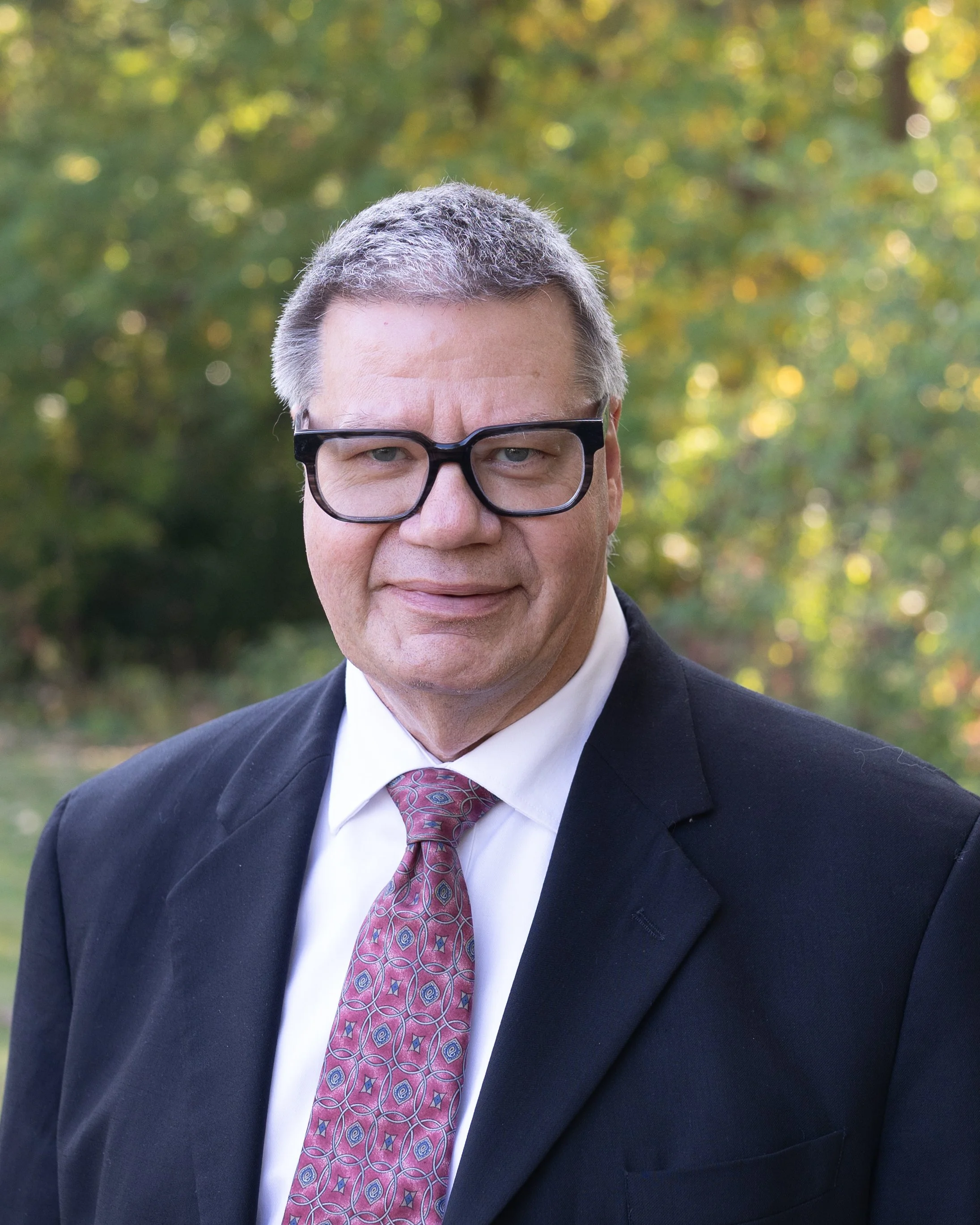 Man wearing glasses, a dark suit jacket with a white shirt, and a red paisley tie, standing outdoors with foliage in the background.