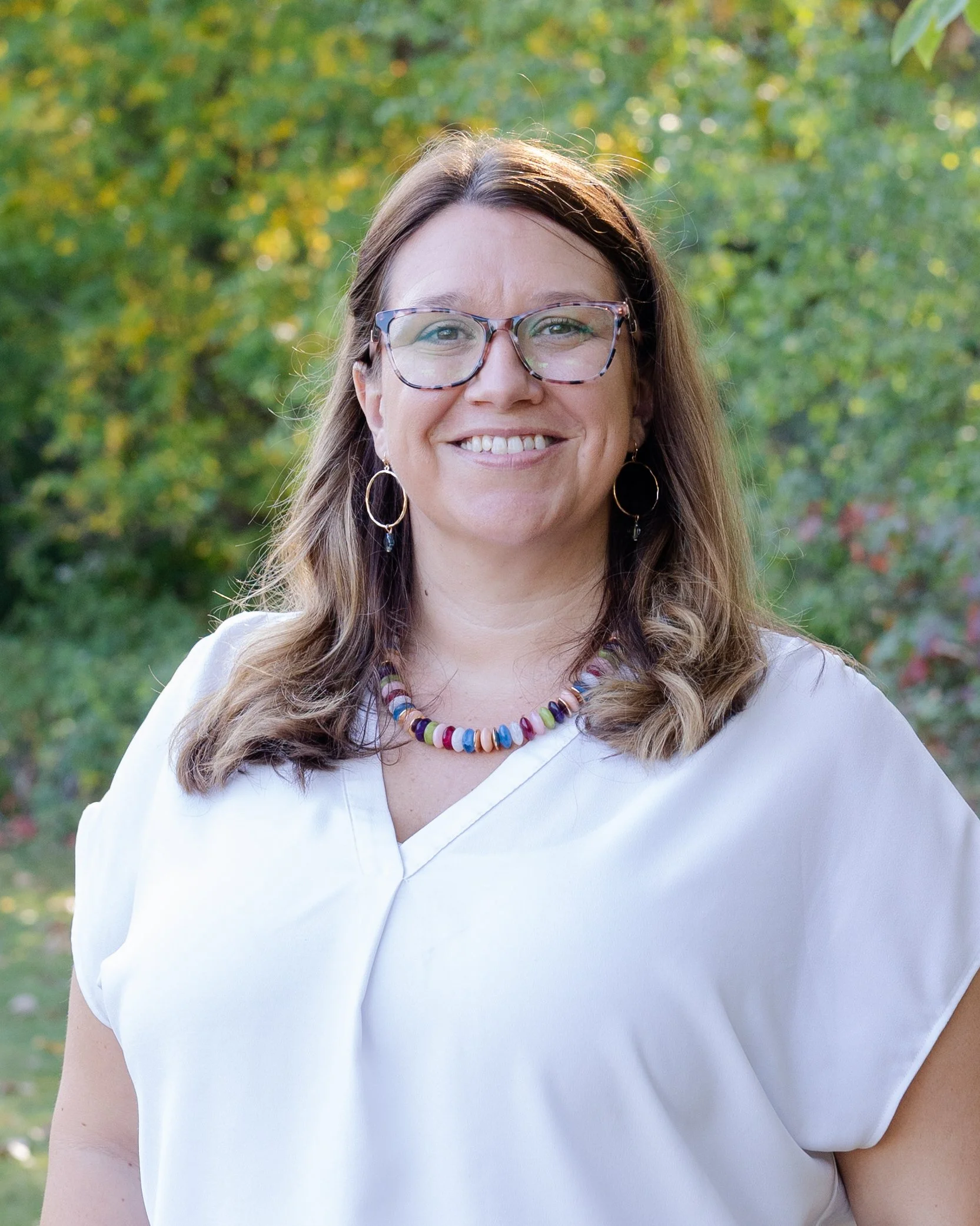 A woman with shoulder-length brown hair, wearing a white shirt and a necklace, smiling outdoors, with green trees in the background.