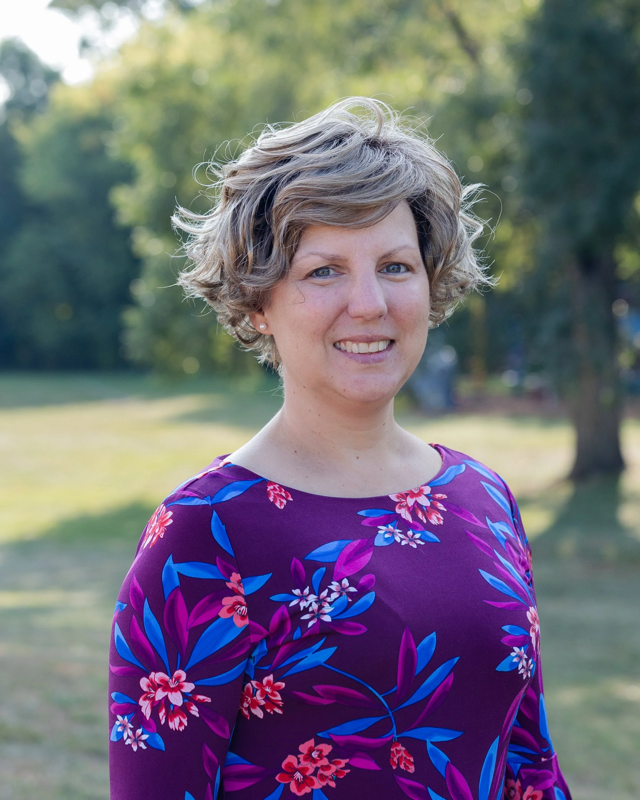 A smiling woman with short blond hair and wearing a patterned top outdoors with a background of green and red foliage.
