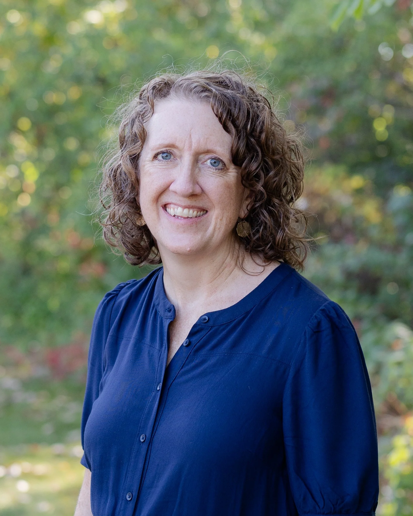 A woman with short, curly brown hair, wearing a denim jacket and a navy blue shirt, smiling outdoors with colorful fall foliage in the background.