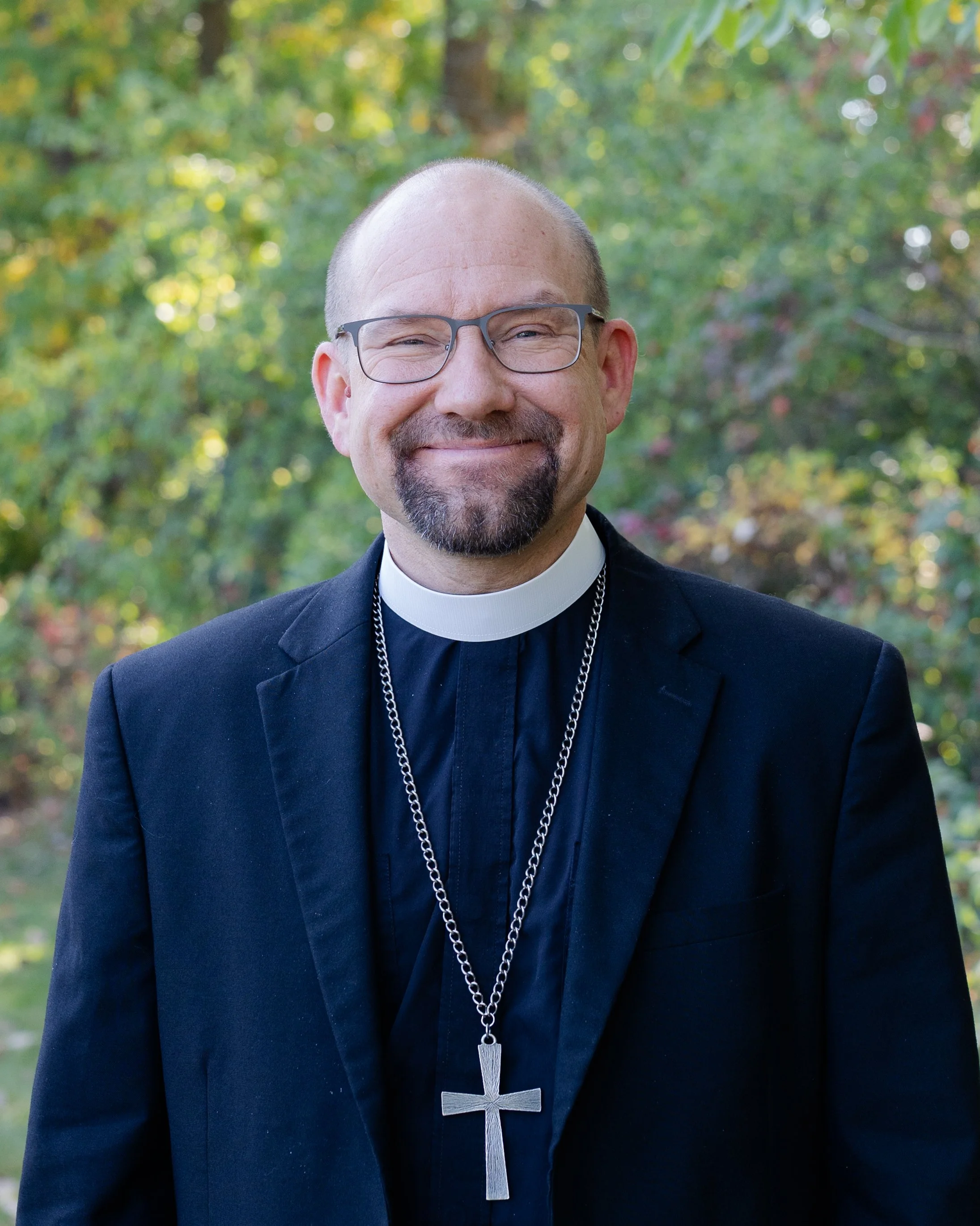 Headshot of a smiling man wearing clerical attire with a black suit and a white clerical collar, standing outdoors with a background of green and red foliage.
