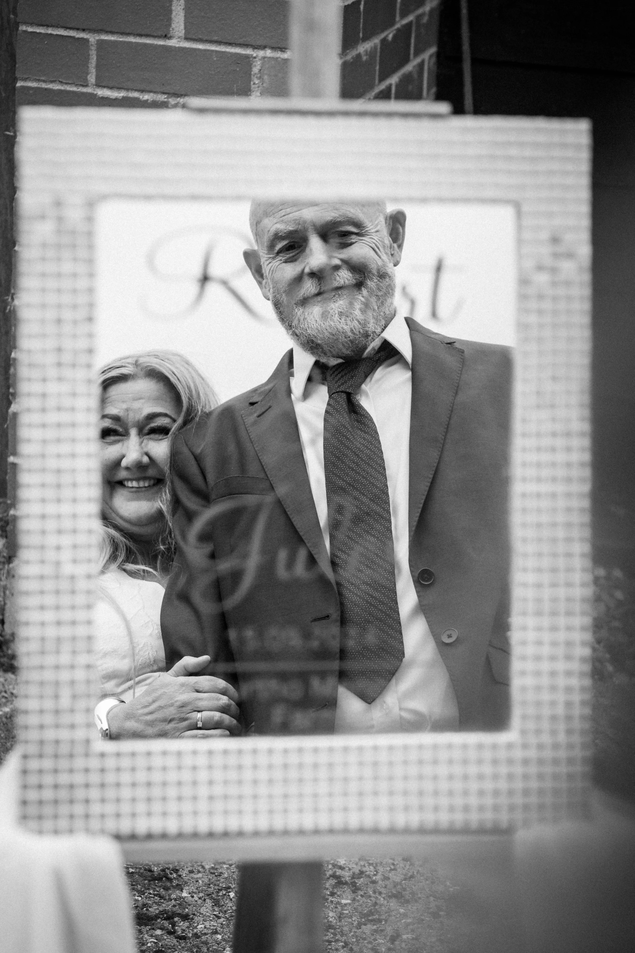 A reflection of a smiling middle-aged man with a beard and a woman, both dressed in formal attire, seen in a mirror or glass surface at a social or celebratory event.