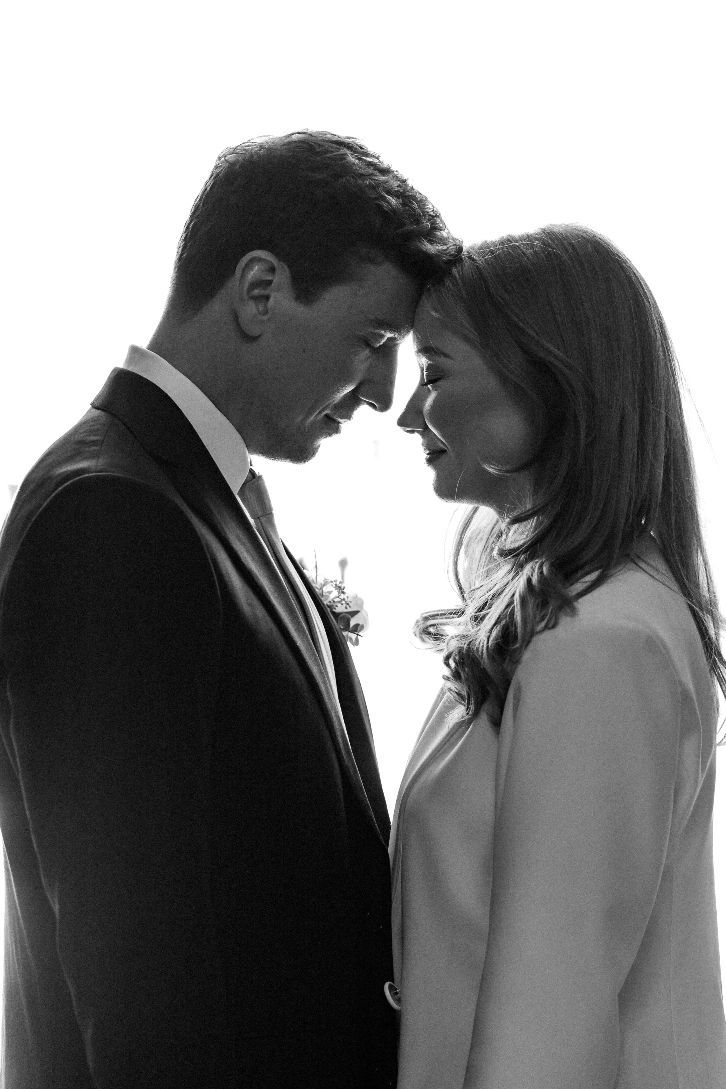 A black and white photo of a man and woman with their foreheads touching, smiling softly at each other, dressed in formal attire, in a close and intimate pose.