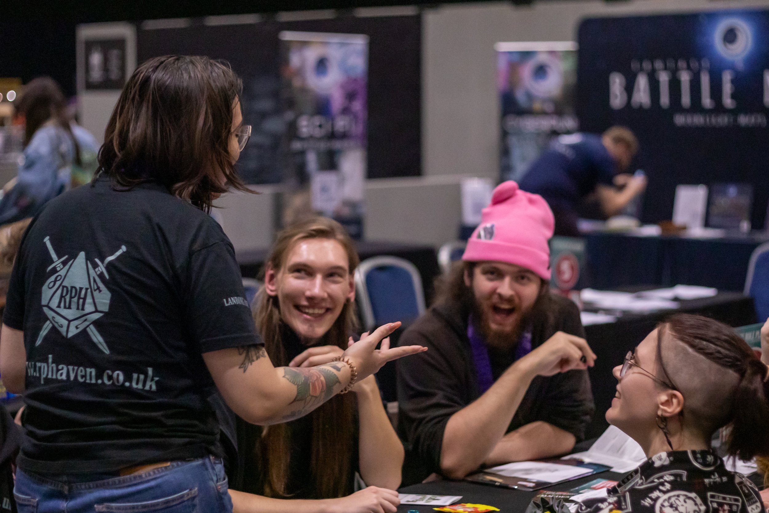 People at a tabletop gaming convention, seated with materials, smiling and talking.