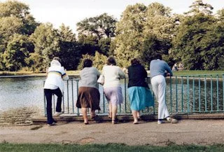 Five people leaning on a railing, looking at a pond with trees in the background.