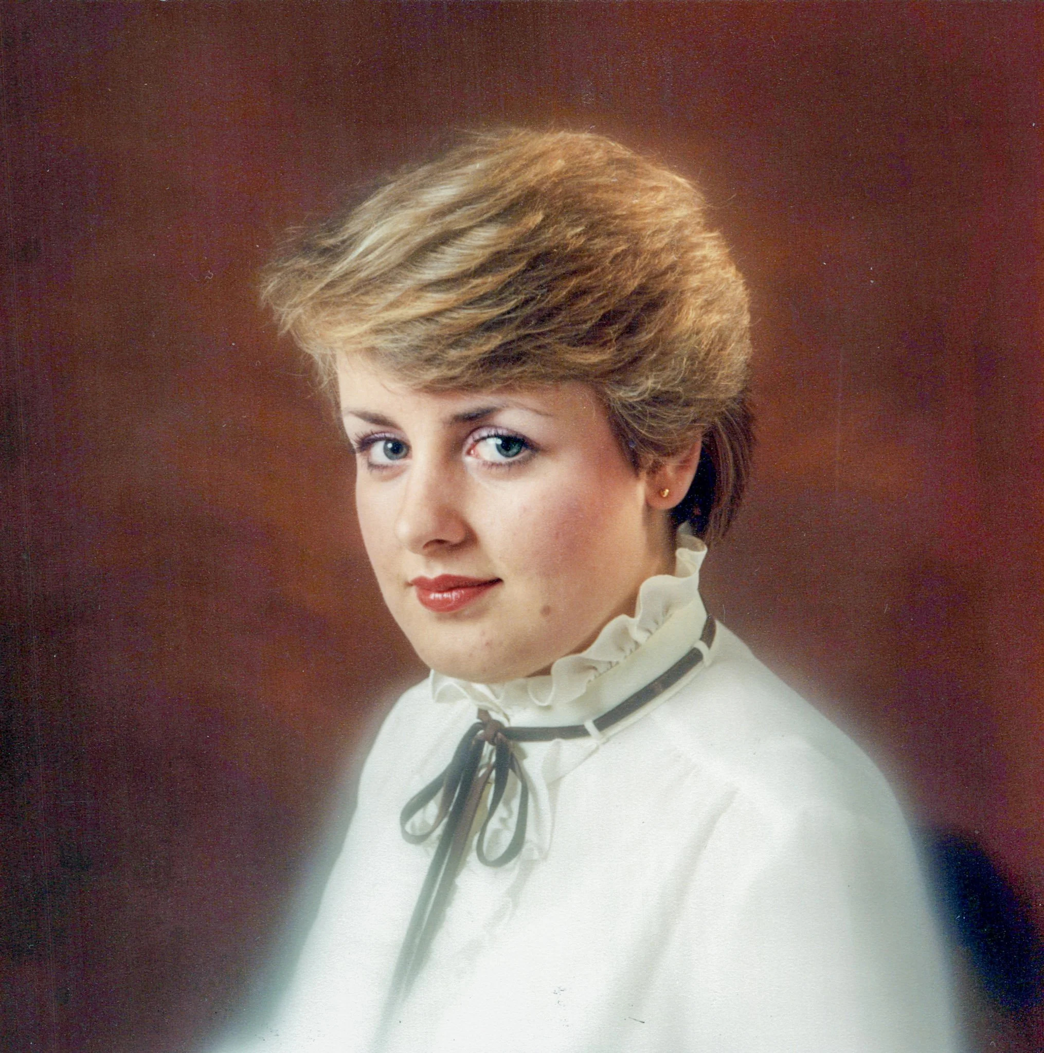 Portrait of a woman with short, styled blonde hair, blue eyes, wearing a white blouse with a ruffled collar and a black ribbon tie, against a dark background.