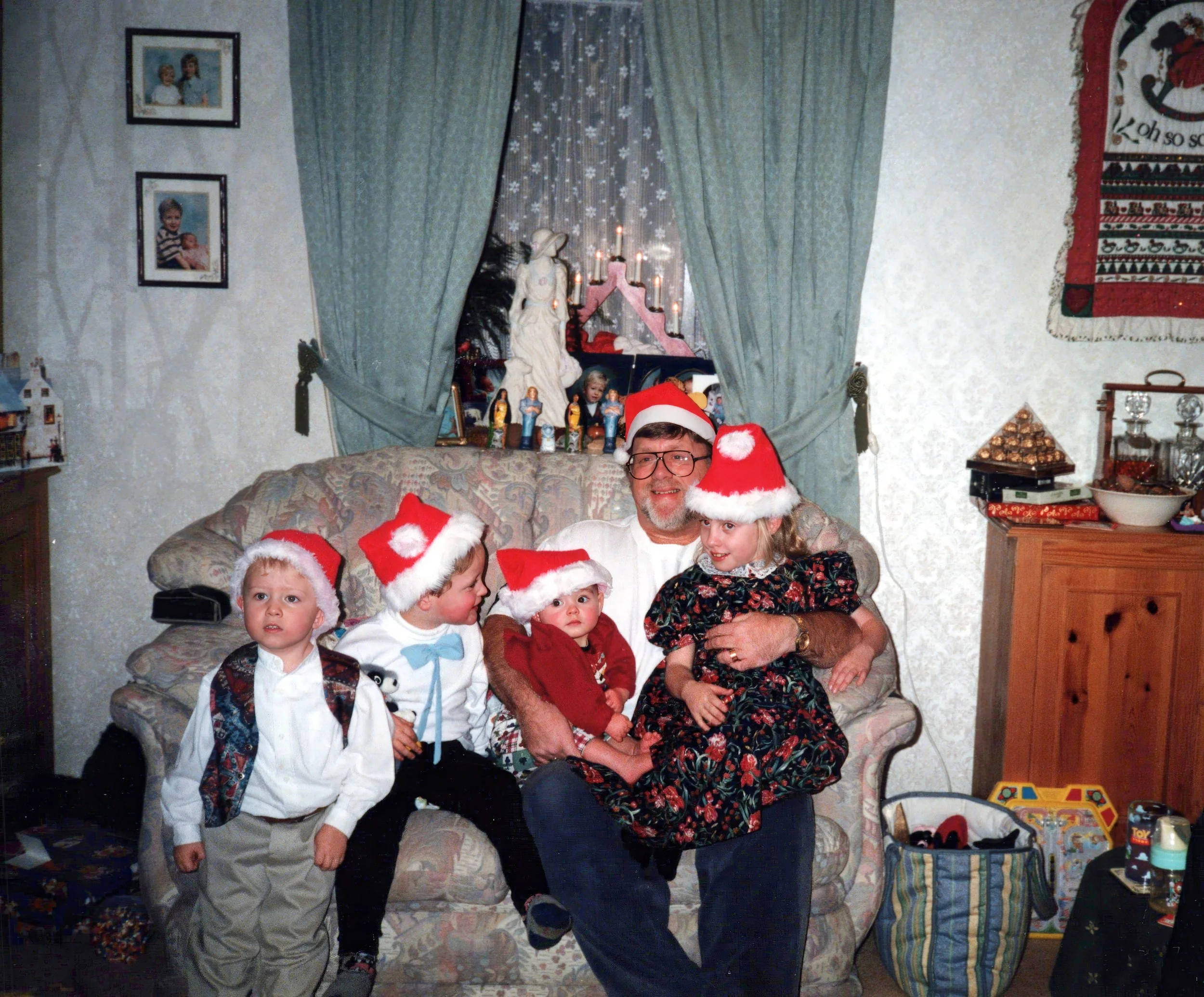 A man wearing glasses and a Santa hat is sitting on a floral-patterned couch, surrounded by four young children also wearing Santa hats. The room is decorated with Christmas ornaments and a nativity scene is visible in the background. The children are of varying ages, dressed in festive clothing. The scene conveys a warm, holiday atmosphere.