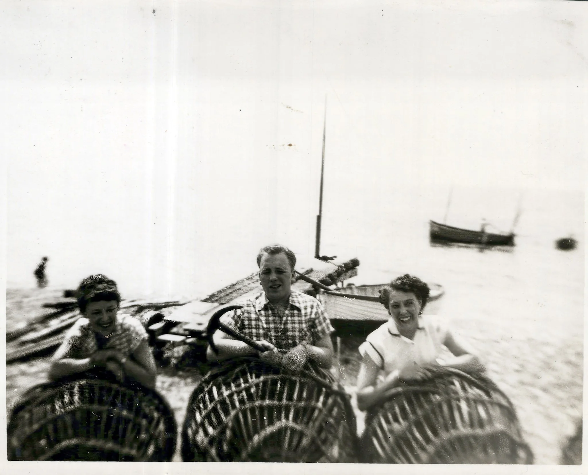 Black and white photo of three smiling people holding large wicker baskets near a beach with boats in the water.