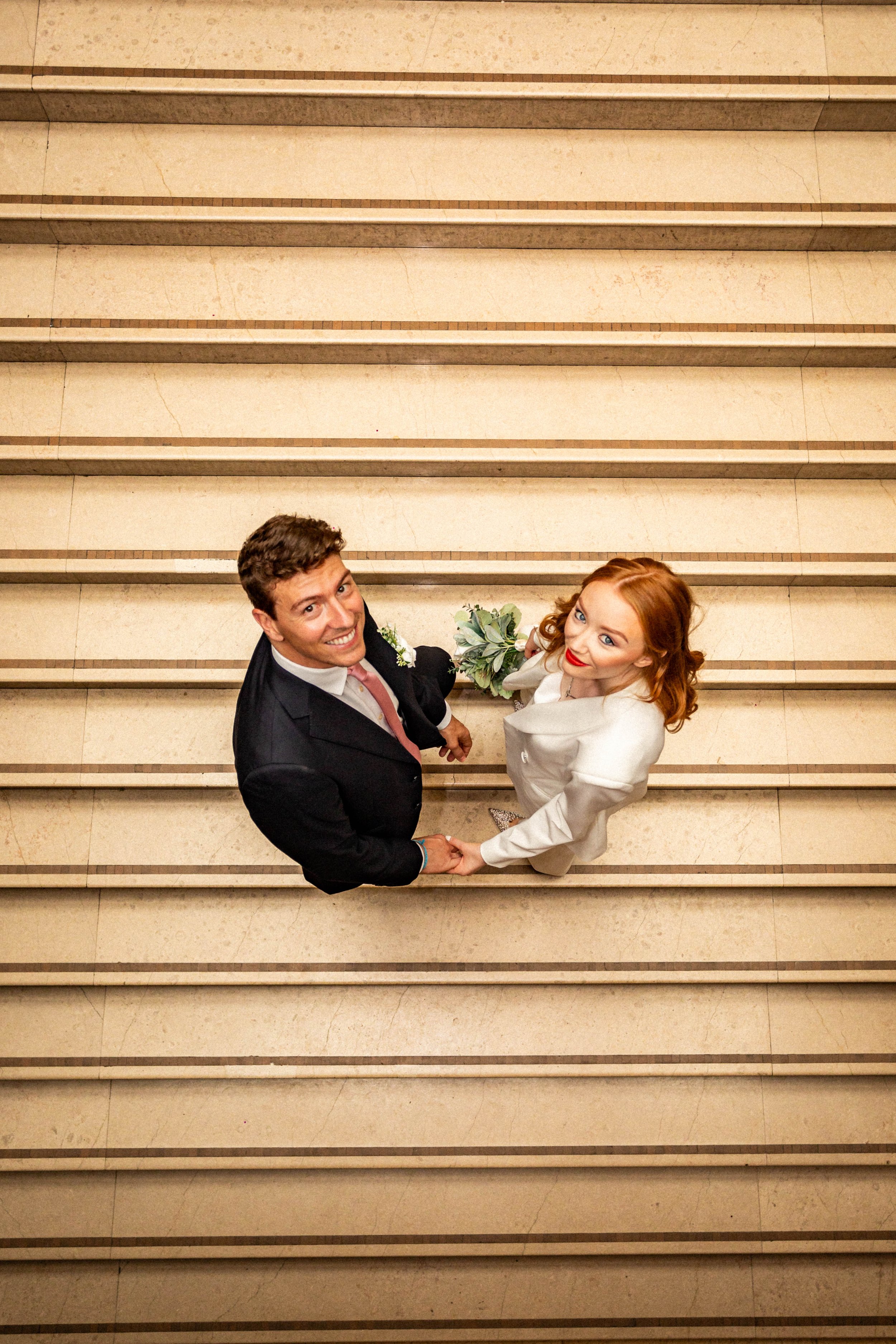 A smiling man and woman in wedding attire holding hands and looking up at the camera on a staircase.