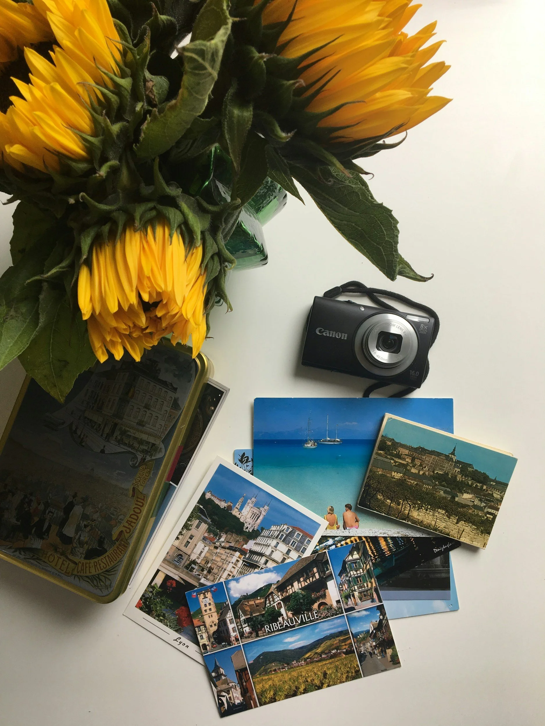 A tabletop with a bouquet of sunflowers, a camera, and several travel postcards featuring scenic views of coastal and city landscapes.