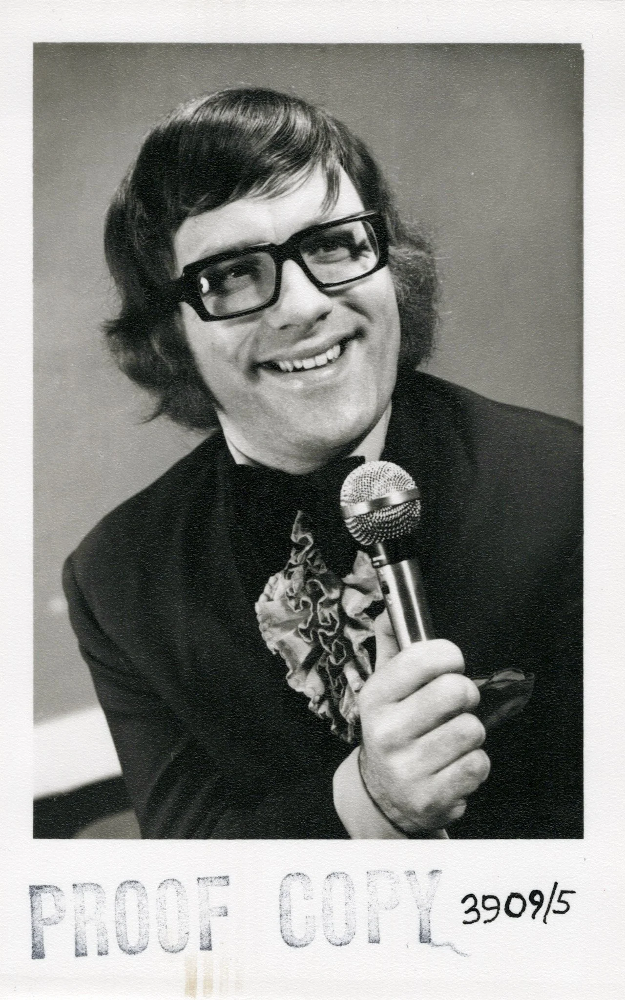 Black and white photo of a man with medium-length hair, glasses, smiling, holding a microphone, wearing a tuxedo with a ruffled shirt.