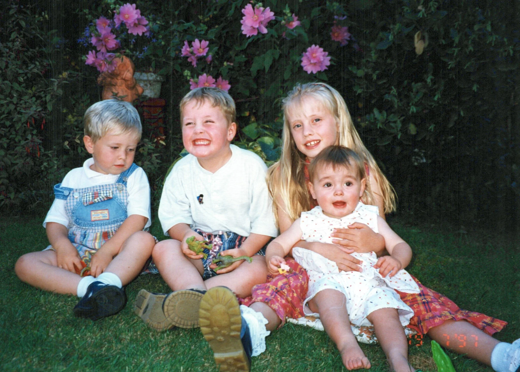 Four children sitting outdoors on grass in front of pink flowers, with two boys and two girls, one young and one older, dressed in summer clothes.