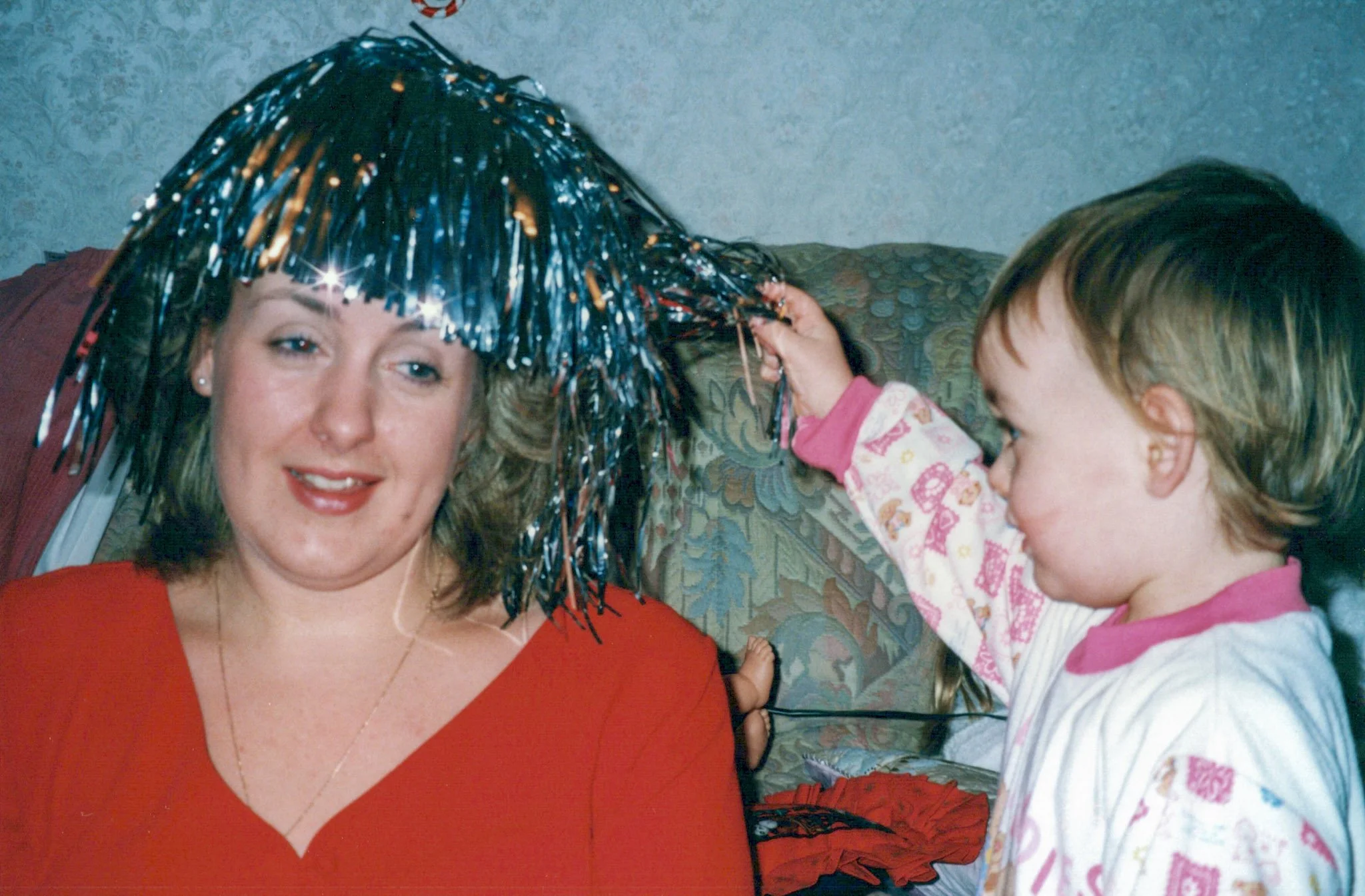 A woman with shoulder-length hair and a red top is being playfully decorated with a shiny blue and silver tinsel hat by a young girl in pajamas, in a living room with a patterned sofa.