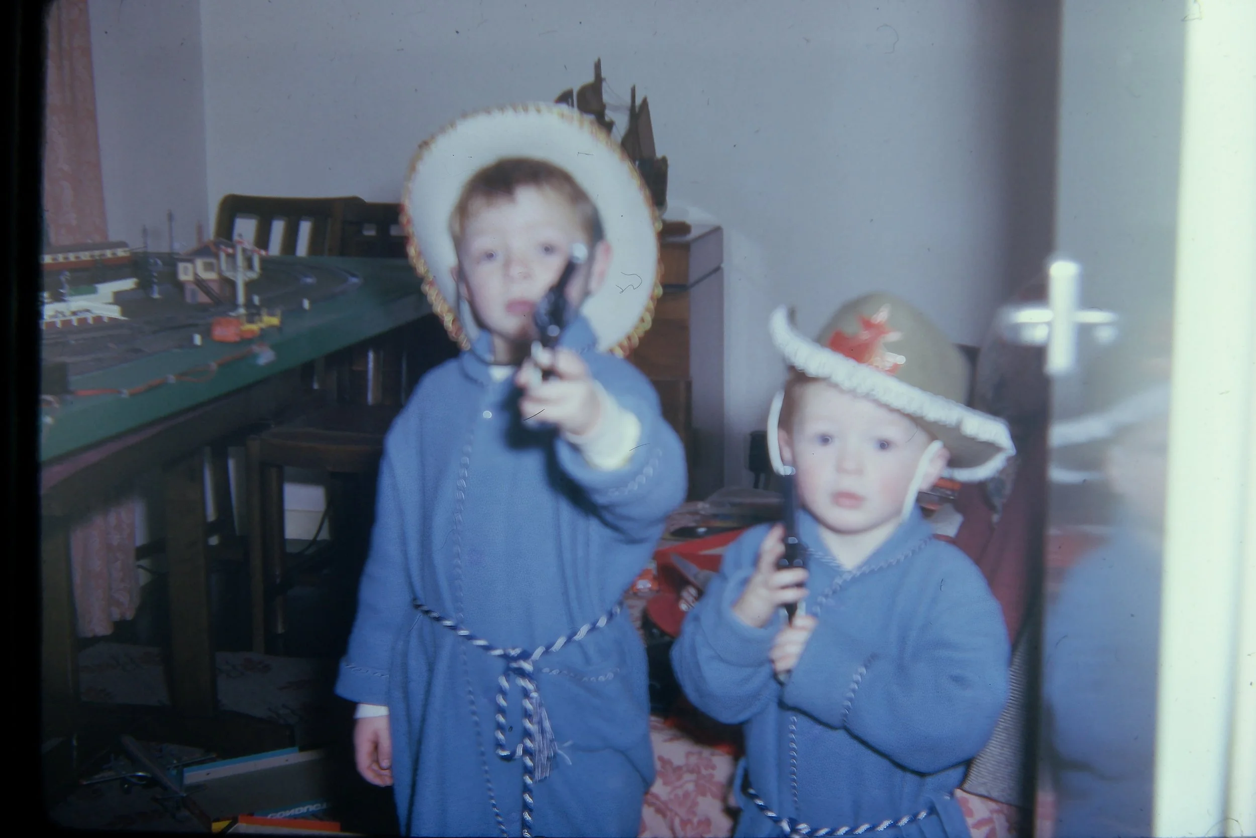 Two young children dressed in blue robes and cowboy hats playing with toy guns indoors. A model train set is visible on a table in the background.