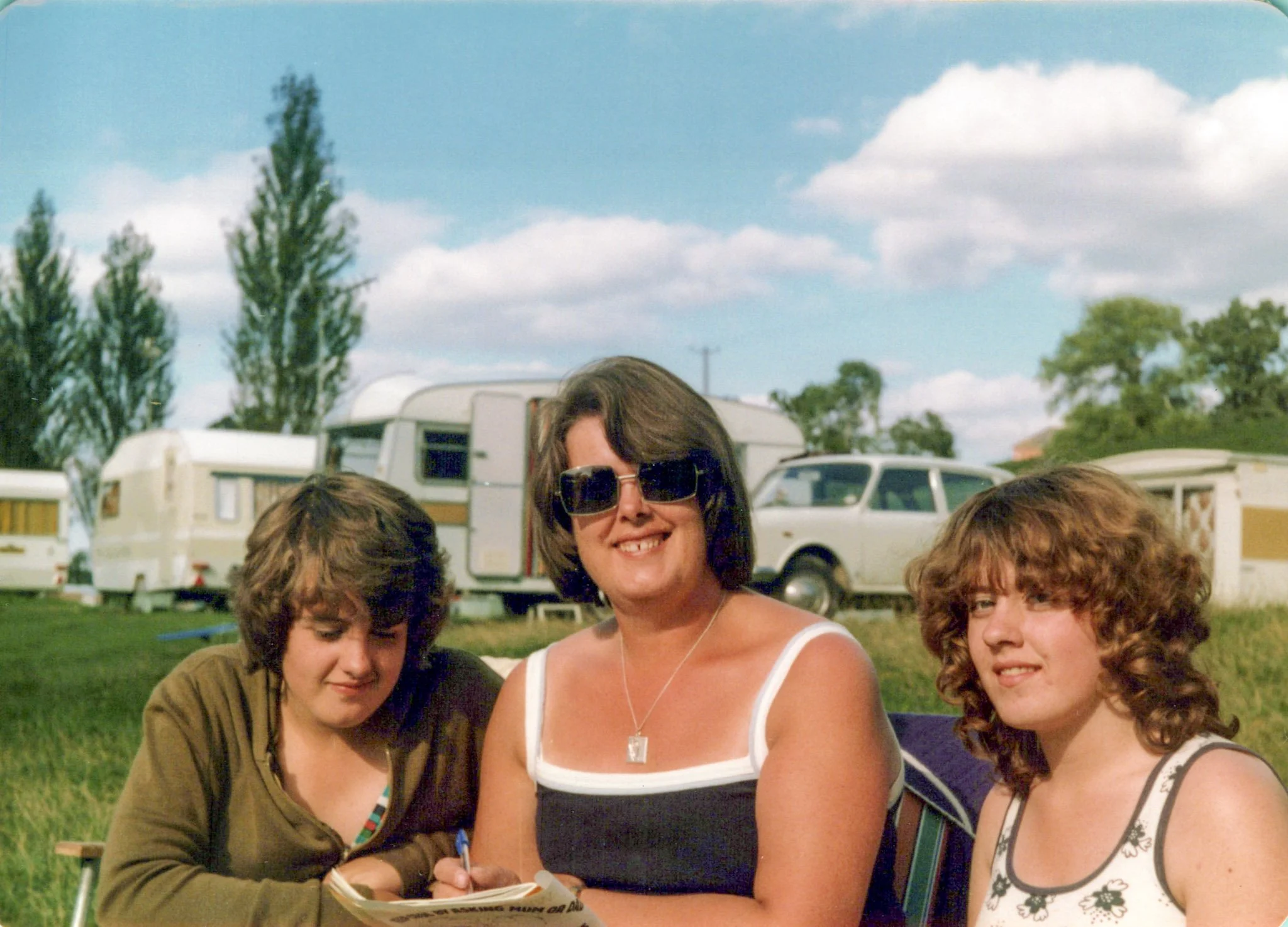 Three women sitting outdoors in a grassy area with caravans, a pickup truck, and trees in the background, on a sunny day with a blue sky and scattered clouds.