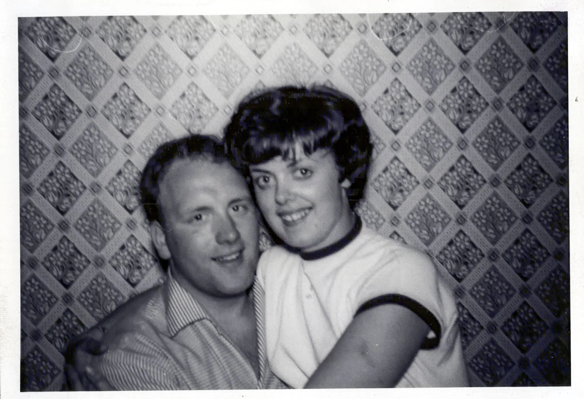 Black and white photo of a man and woman smiling, closely embracing, with a patterned wall in the background.