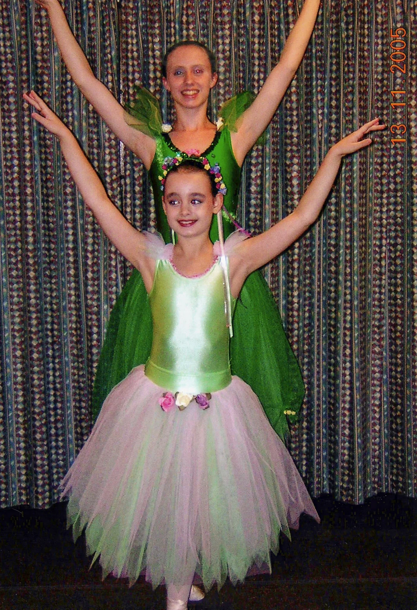 Two young girls dressed in fairy or princess costumes, with wings and tutus, pose with arms raised in front of a patterned curtain.