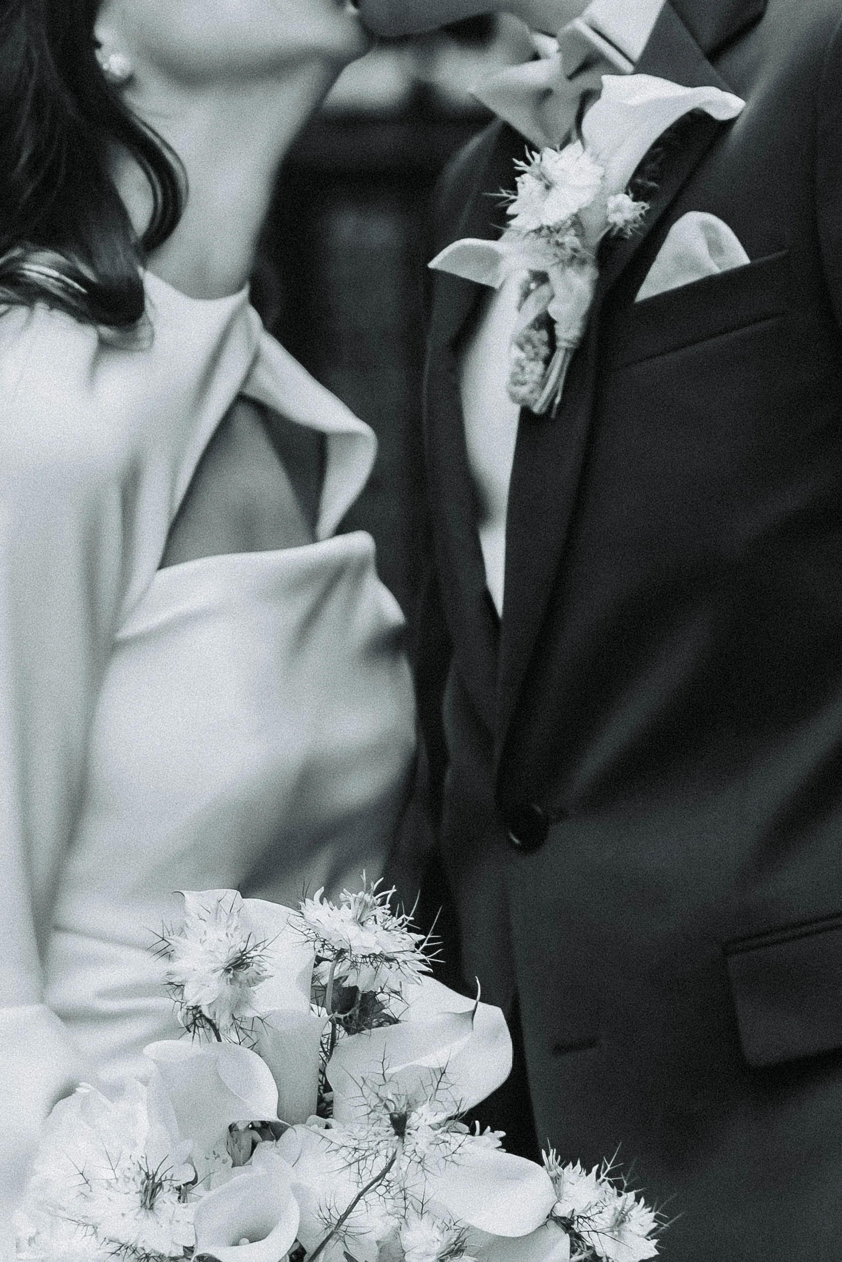 Close-up of a bride and groom standing close, with the bride holding a bouquet of flowers. The groom is wearing a tuxedo with a floral boutonniere, and the bride's shoulder and dress are partially visible. The image is in black and white.