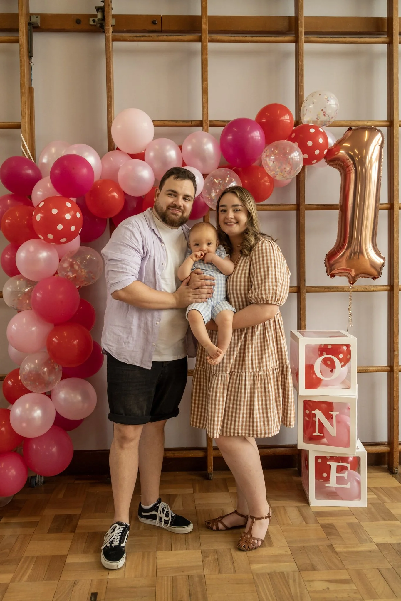 A family of three standing in front of a decorated backdrop with pink, red, and transparent balloons, and a large rose gold number one balloon, celebrating a first birthday. The mother is holding a baby, and the father is standing beside them.